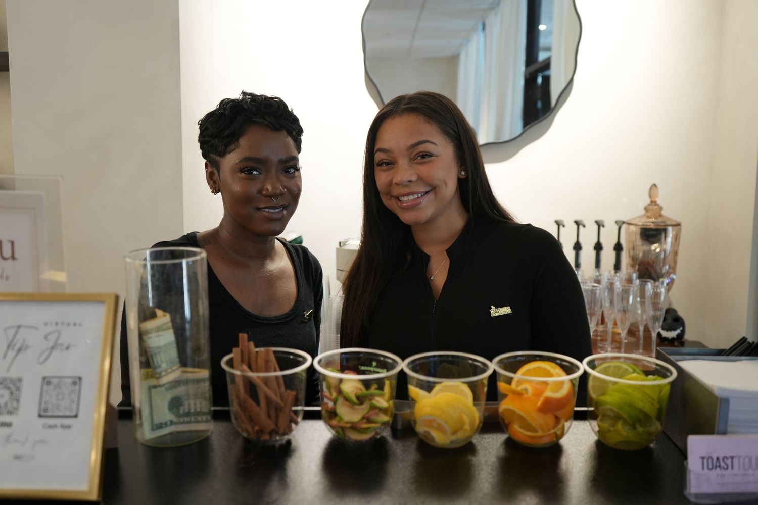Two women smiling behind a bar counter with garnishes, cinnamon sticks, and citrus fruits at a cocktail event.