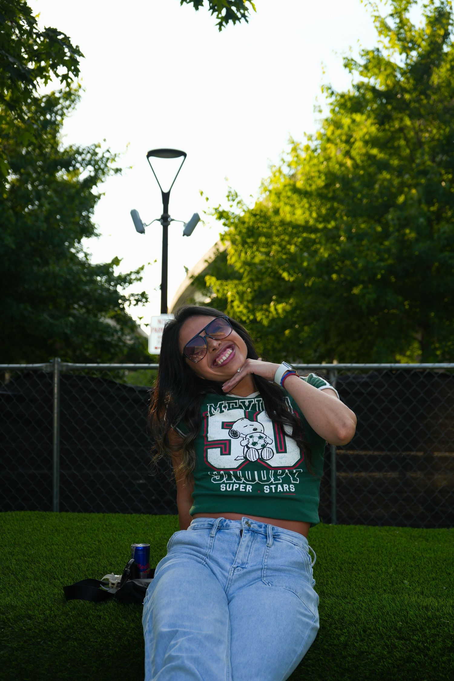 Smiling young woman in green Snoopy shirt and jeans sitting on grass in a park with trees and lamp post.