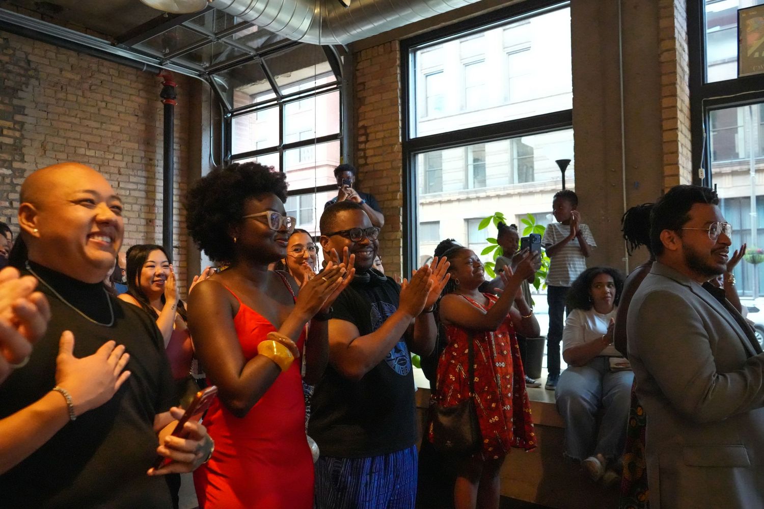 Diverse group of people clapping and smiling at an indoor event in a brick-walled venue with large windows.