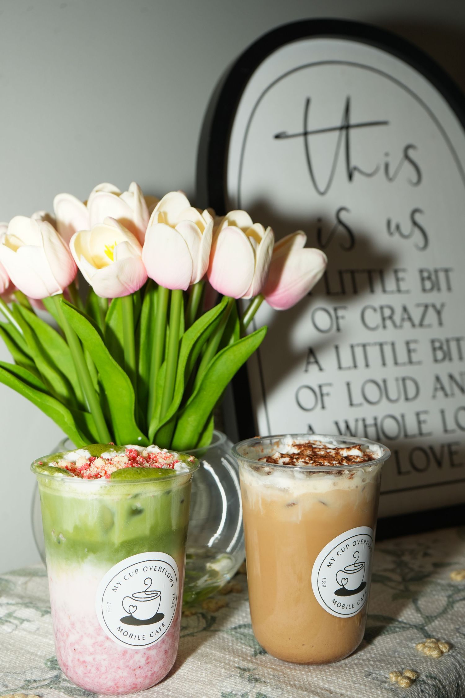 Two iced lattes in glass mugs beside pink tulips, with a decorative sign reading This Is Us in the background.