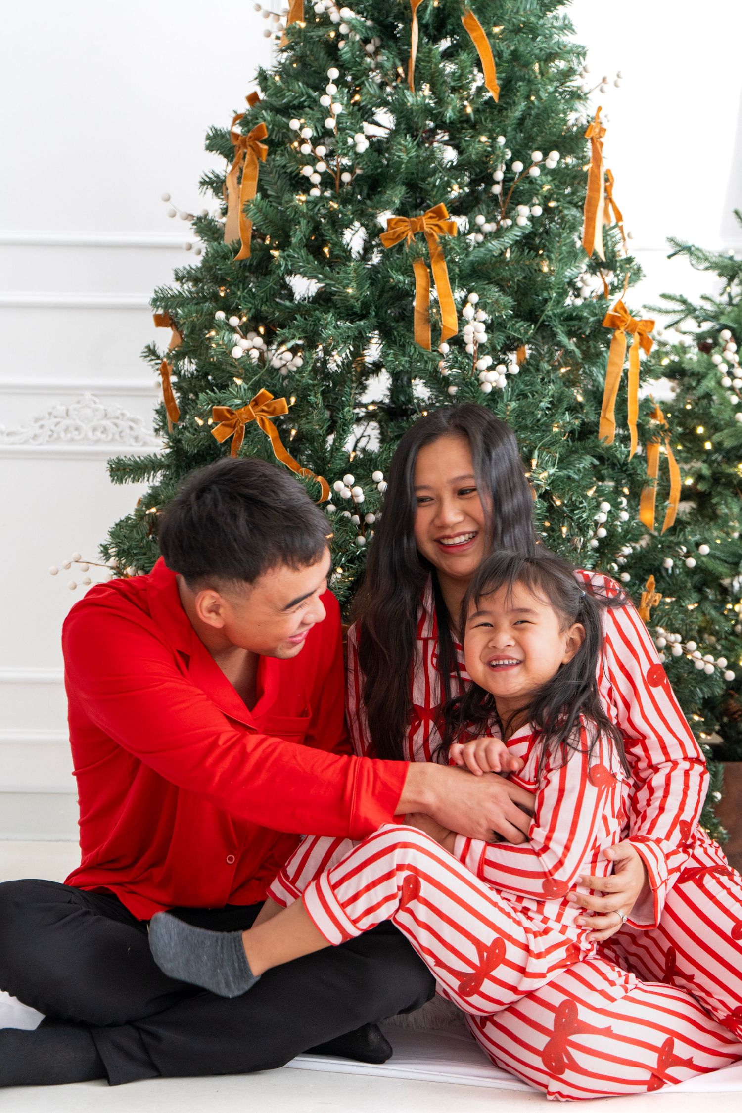 Happy Asian family in matching Christmas pajamas sitting together in front of decorated Christmas tree.
