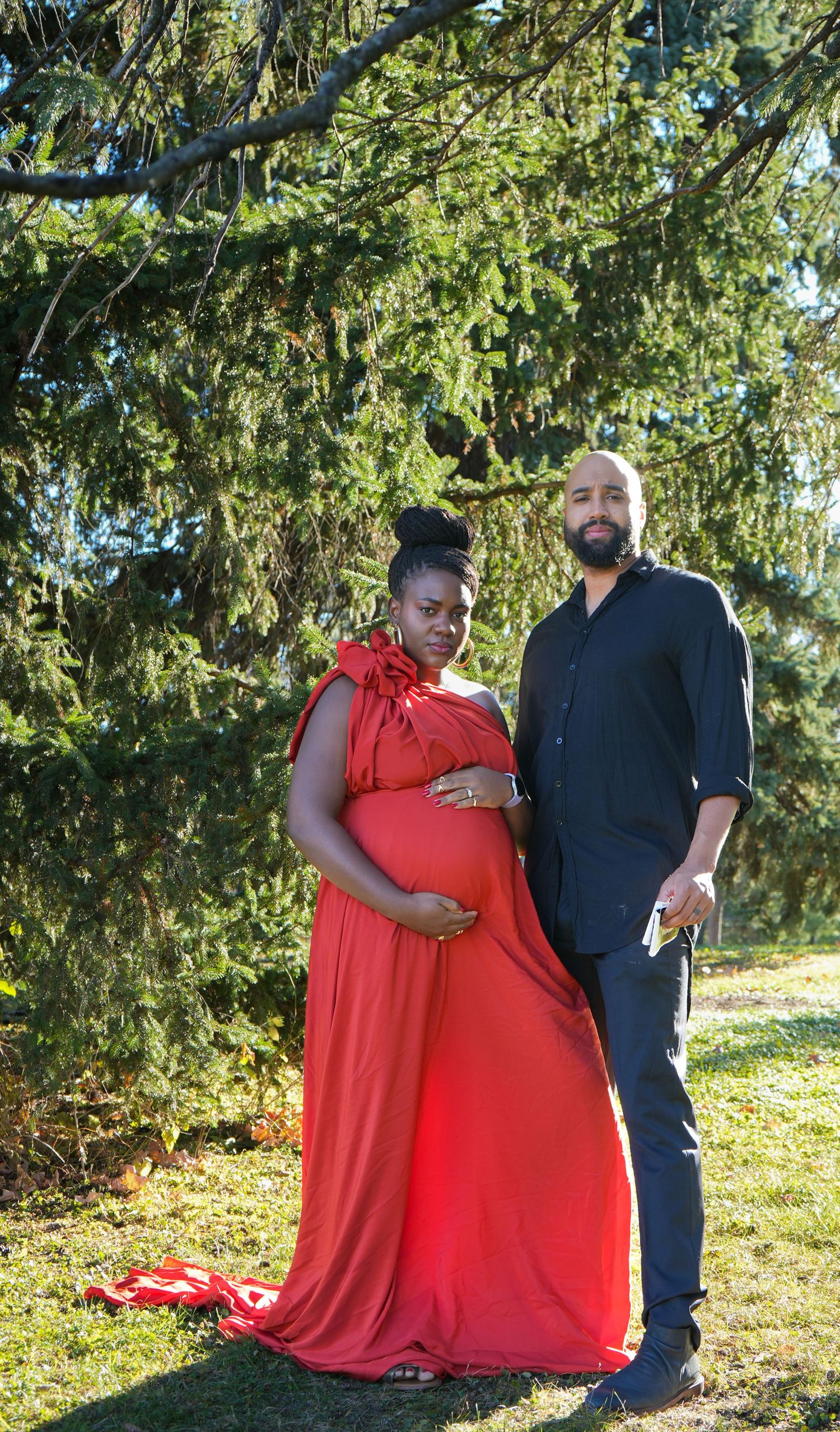 Pregnant woman in red maternity gown poses with bearded man in black beside lush green trees outdoors.