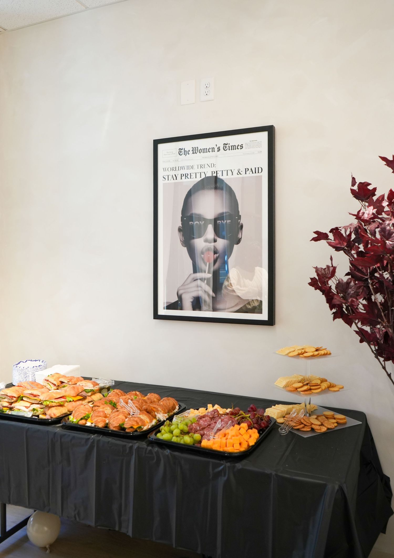 Elegant party spread with charcuterie and snacks on black tablecloth beneath framed Women's Times poster.