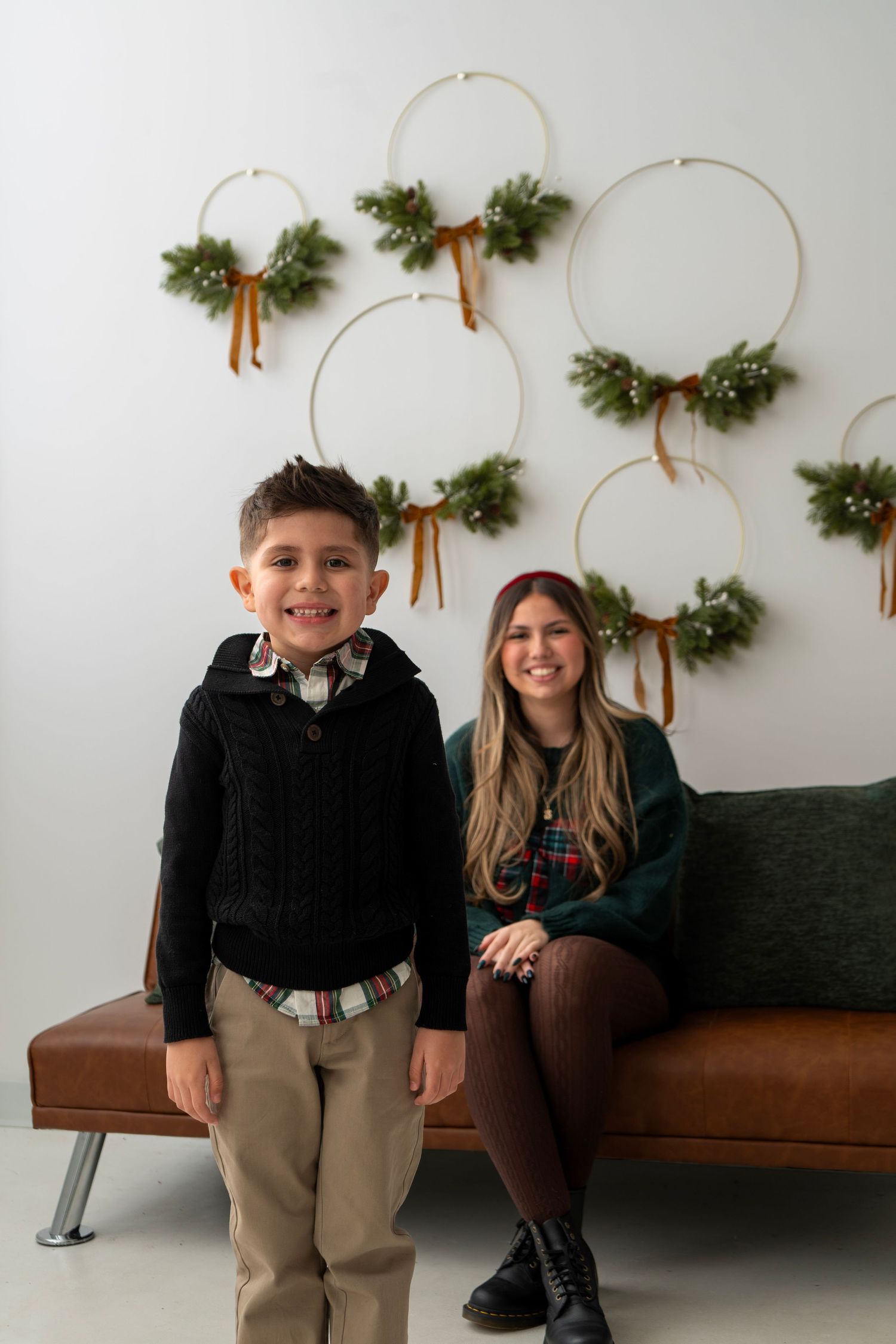 Boy and girl posing for Christmas photo in front of festive hoop wreaths with pine and ribbon on white wall.