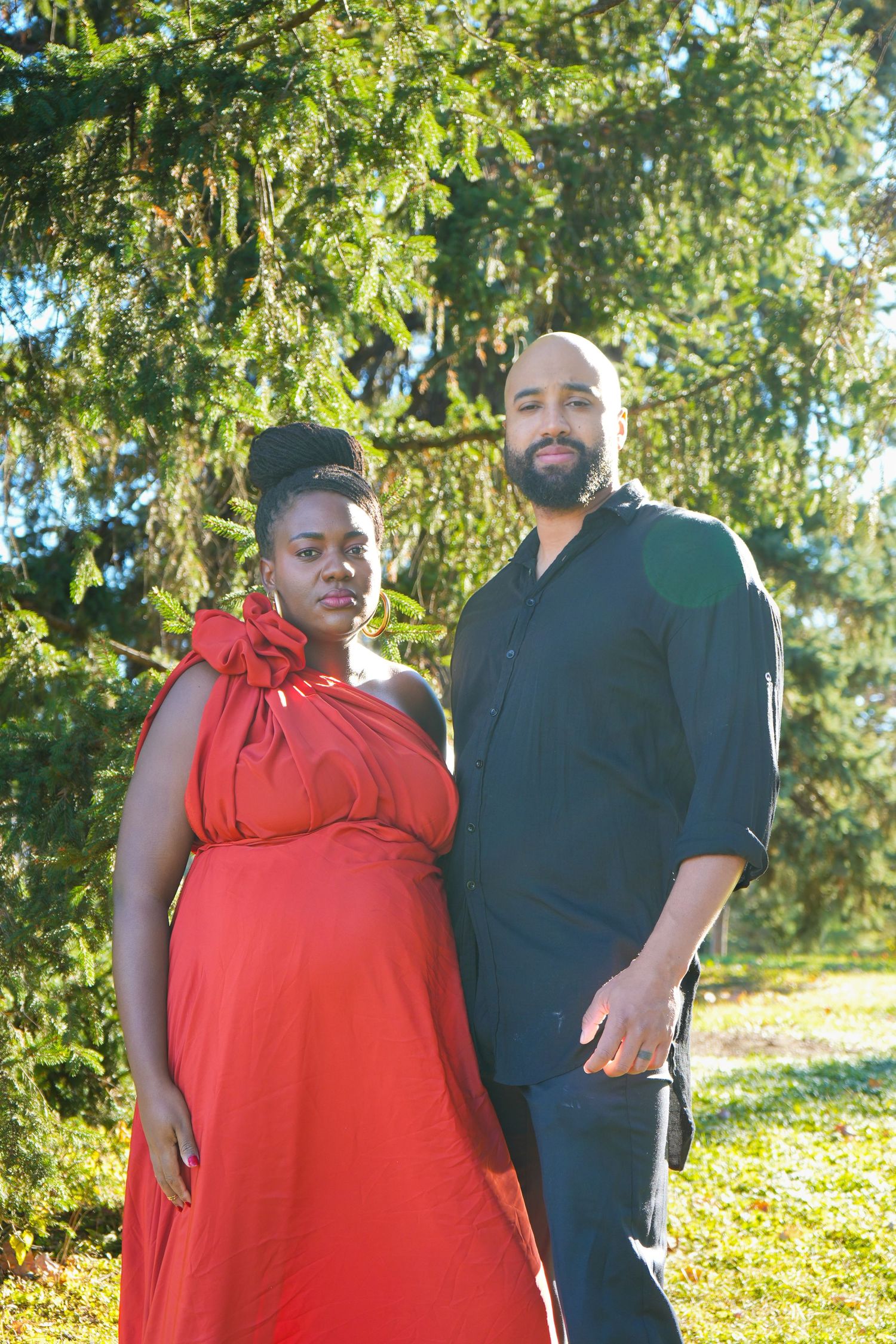 Pregnant woman in red dress and man in black shirt posing outdoors near evergreen trees on a sunny day.