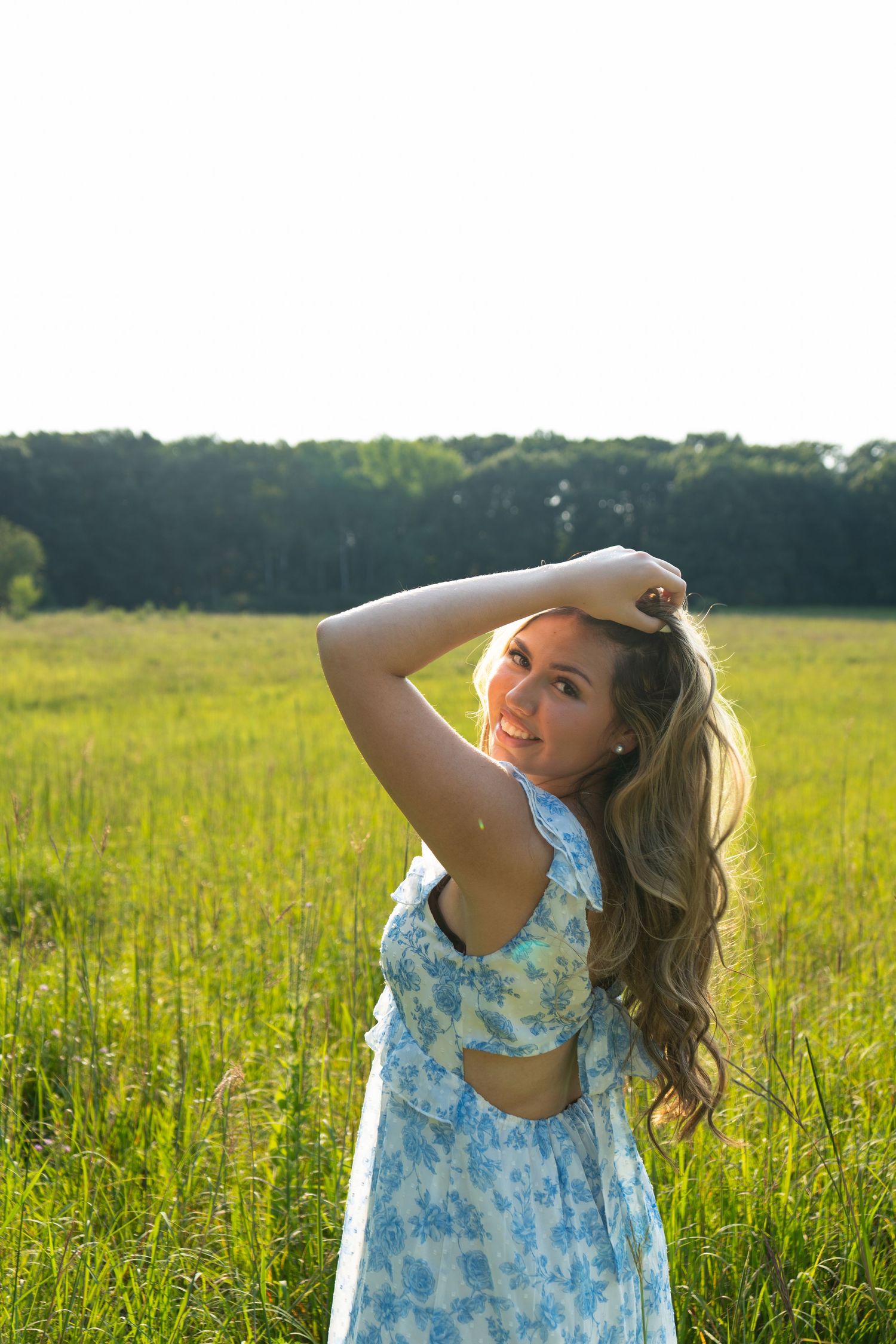 Young woman in blue floral dress standing in sunny green meadow, hand in hair, smiling over shoulder.