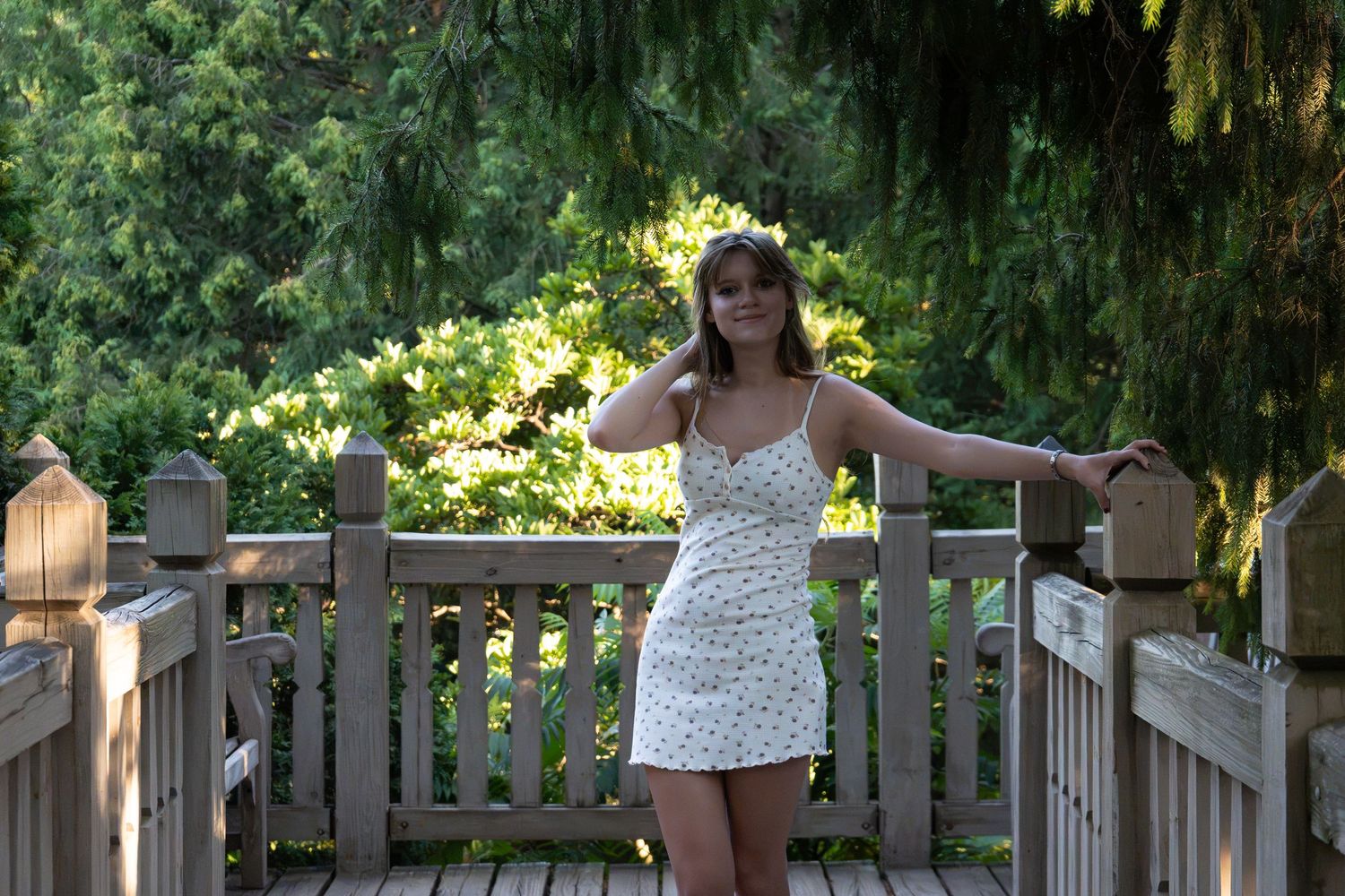 Young woman in floral sundress posing on wooden deck bridge surrounded by lush green trees.