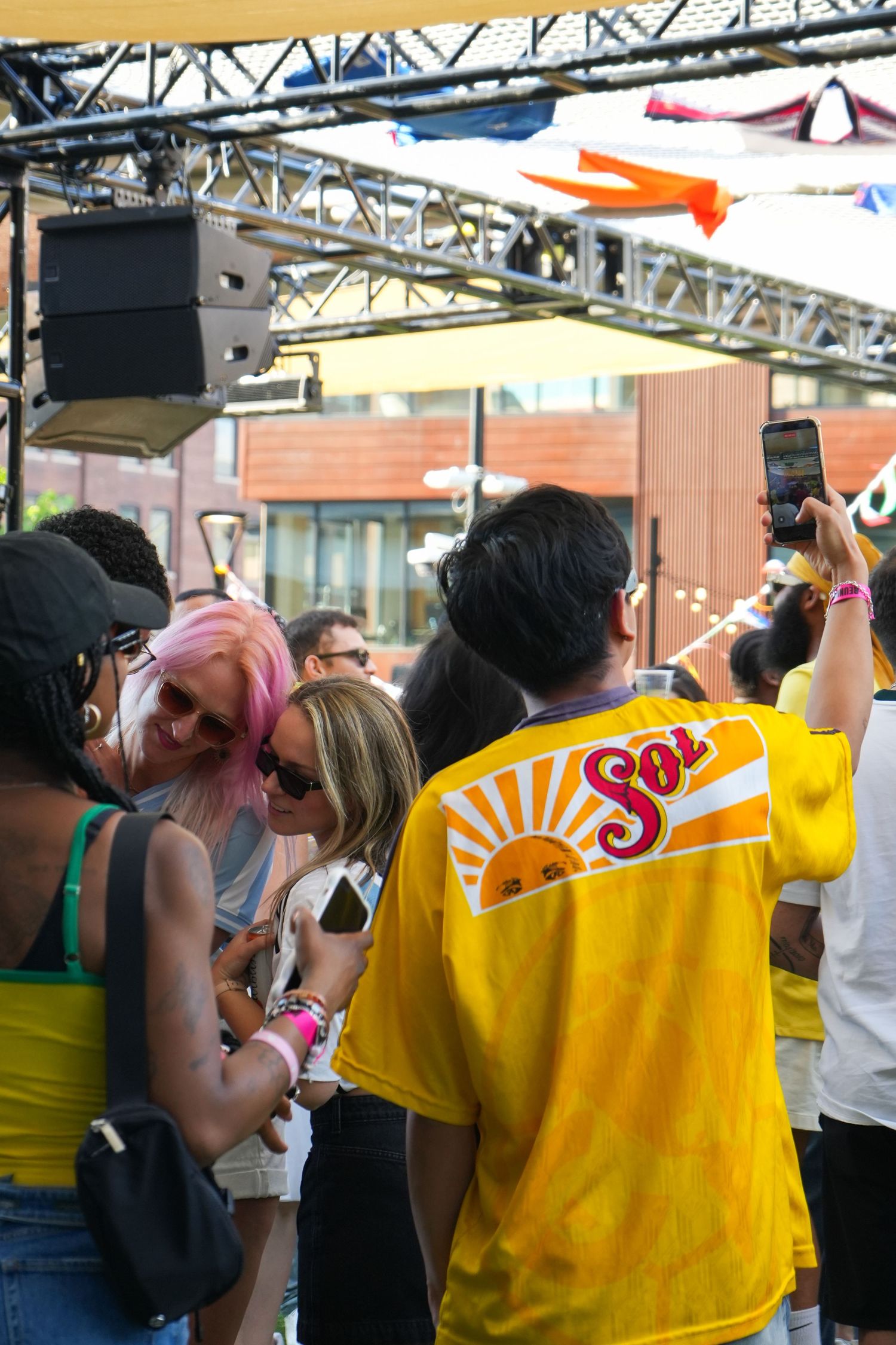 Crowd at outdoor music festival, person in yellow Sol shirt takes phone photo near stage with speakers.