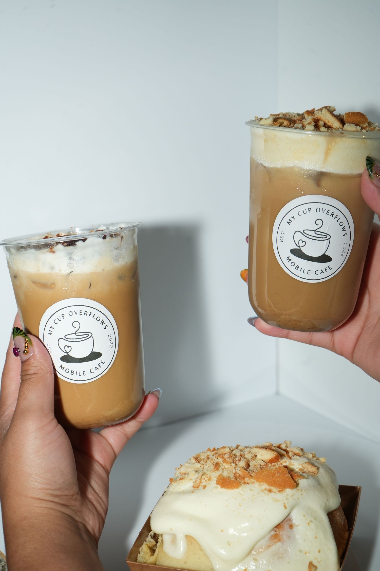 Two hands holding iced coffee drinks from My Cup Overflows Mobile Cafe, with a pastry visible in the foreground.