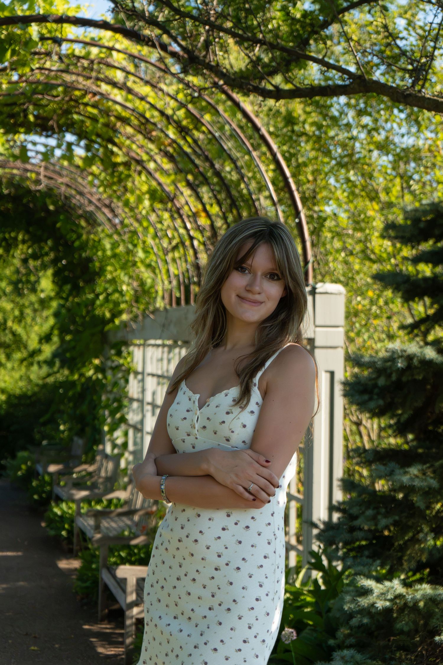 Young woman in floral sundress poses with arms crossed under a lush green garden arbor on a sunny day.