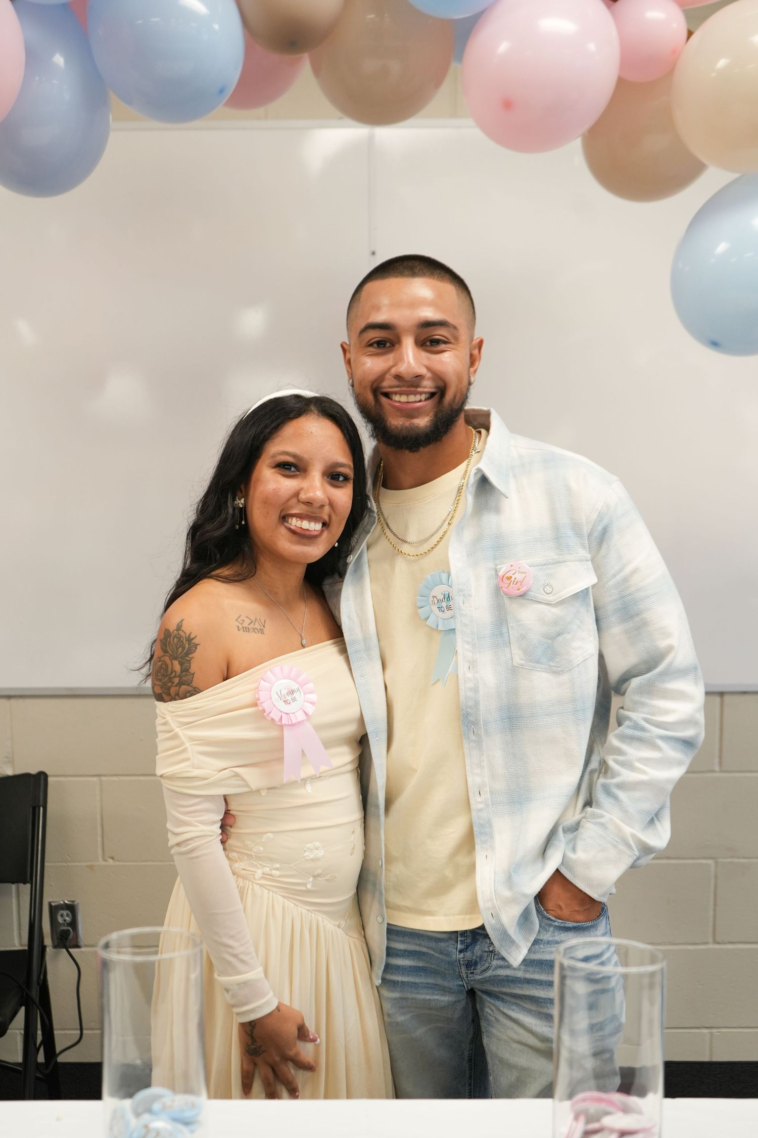 Smiling couple at a gender reveal party with pink and blue balloons, woman in cream dress with pink rosette, man in plaid shirt.