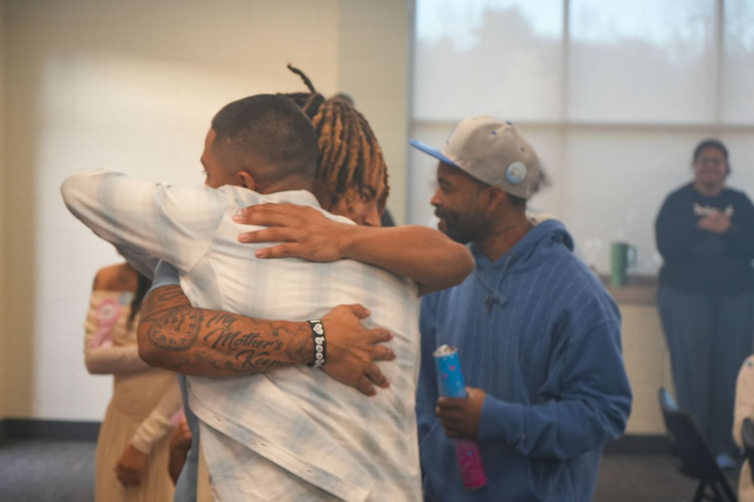 Two men share an emotional hug in a community center while others look on in support.