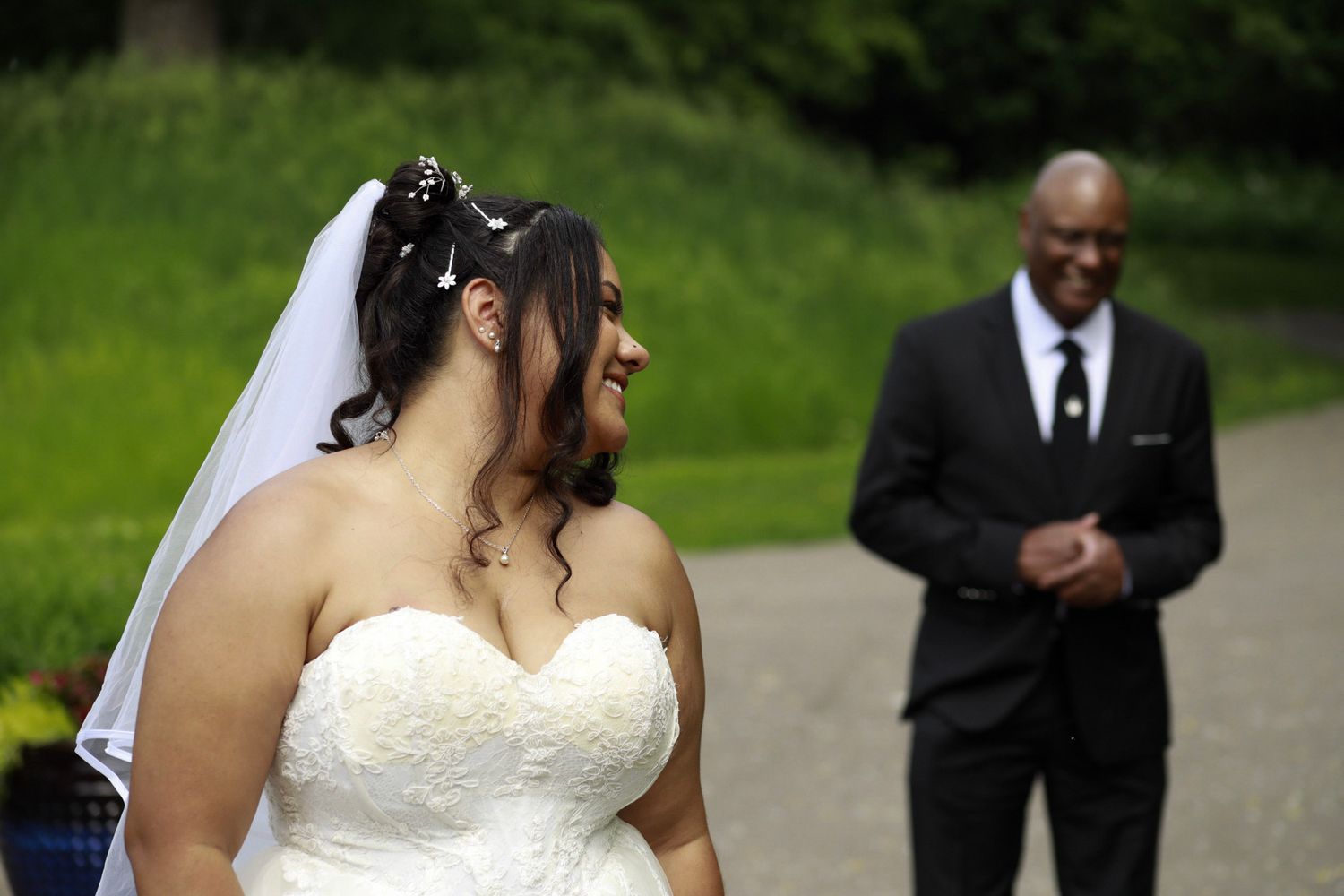 Bride in white strapless gown and veil smiles outdoors, with groom in black suit standing in background.