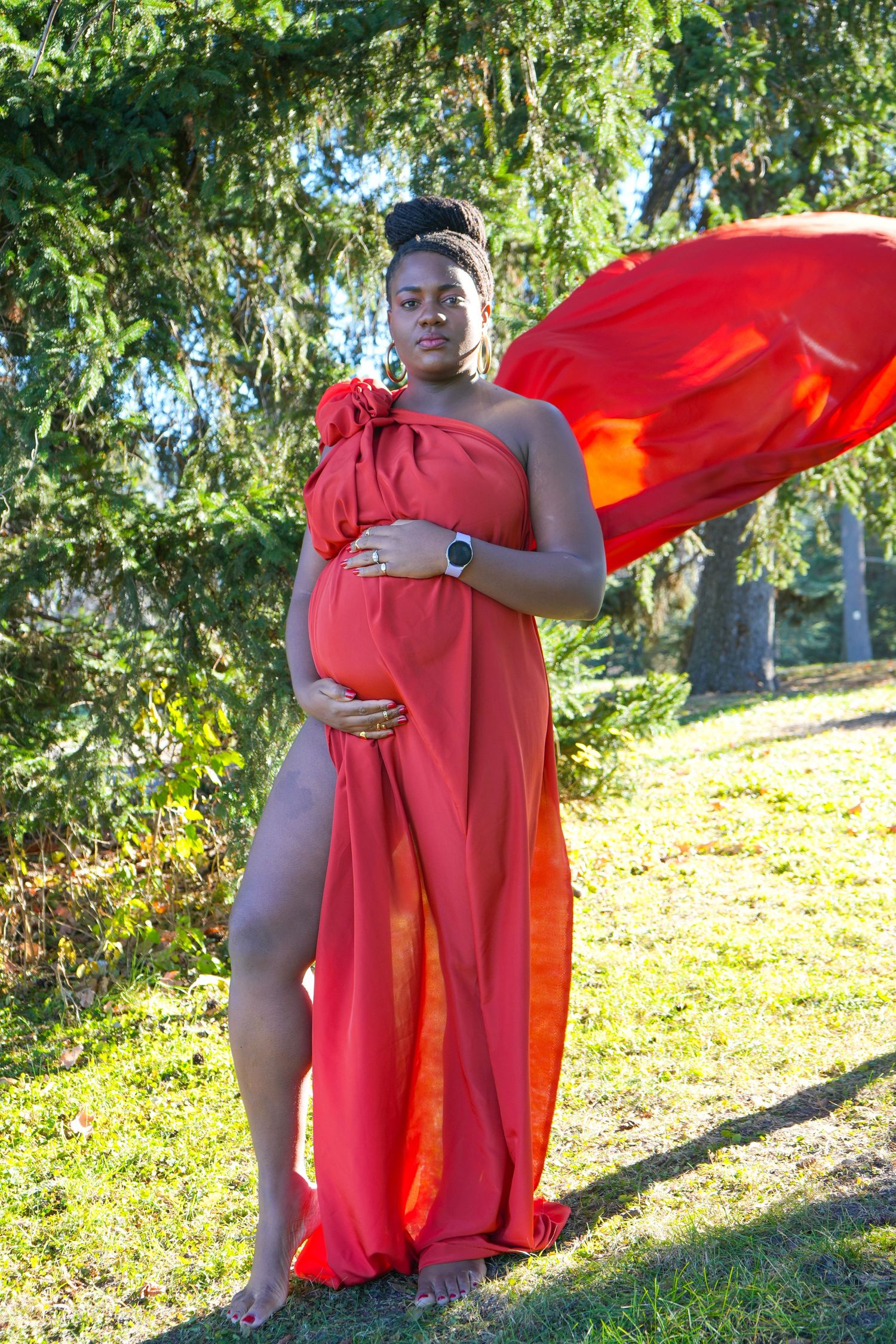 Pregnant woman in flowing red dress poses barefoot outdoors among green pine trees, cradling her baby bump.