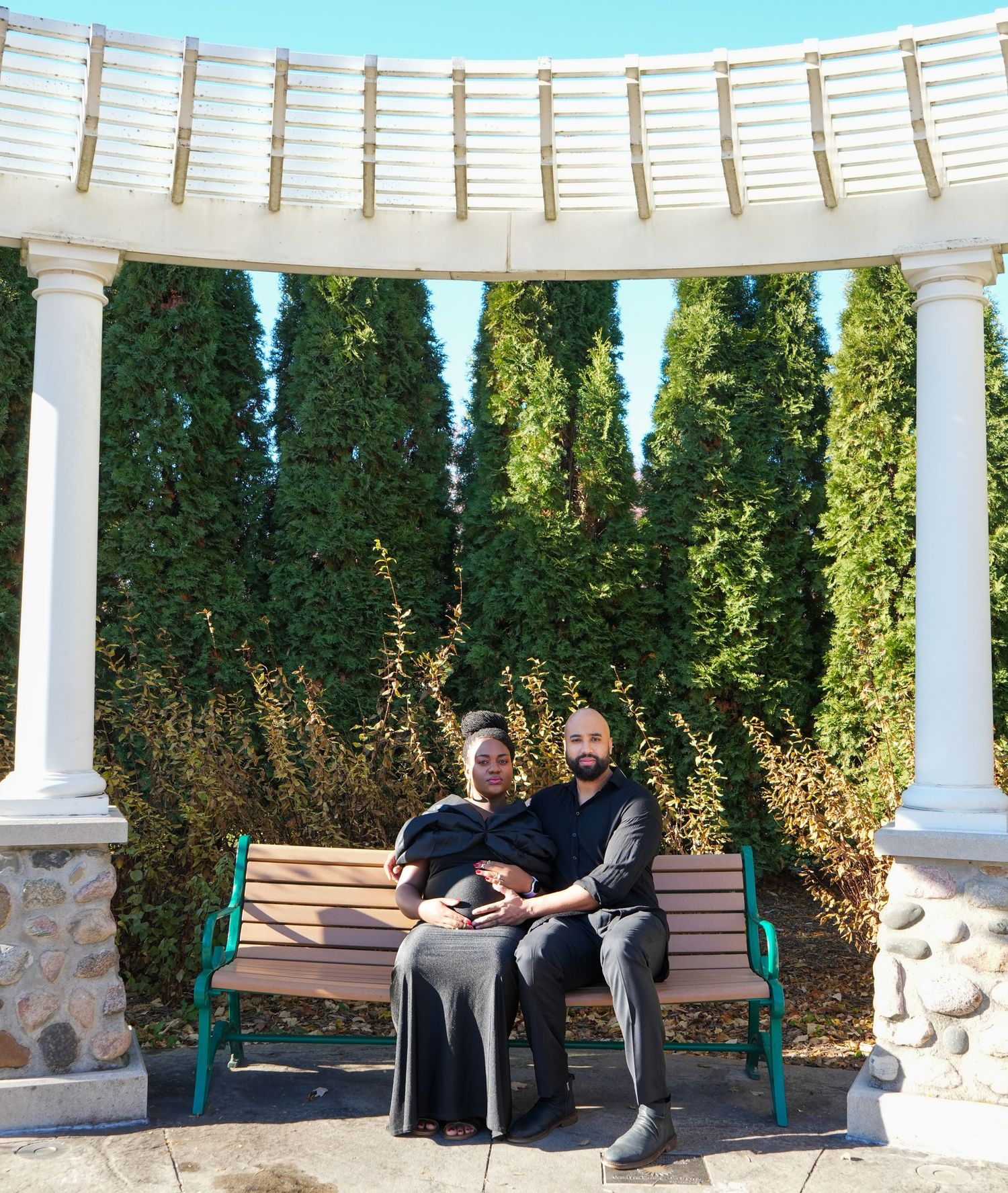 Couple sitting on a bench under a white pergola surrounded by lush green trees in an outdoor garden setting.