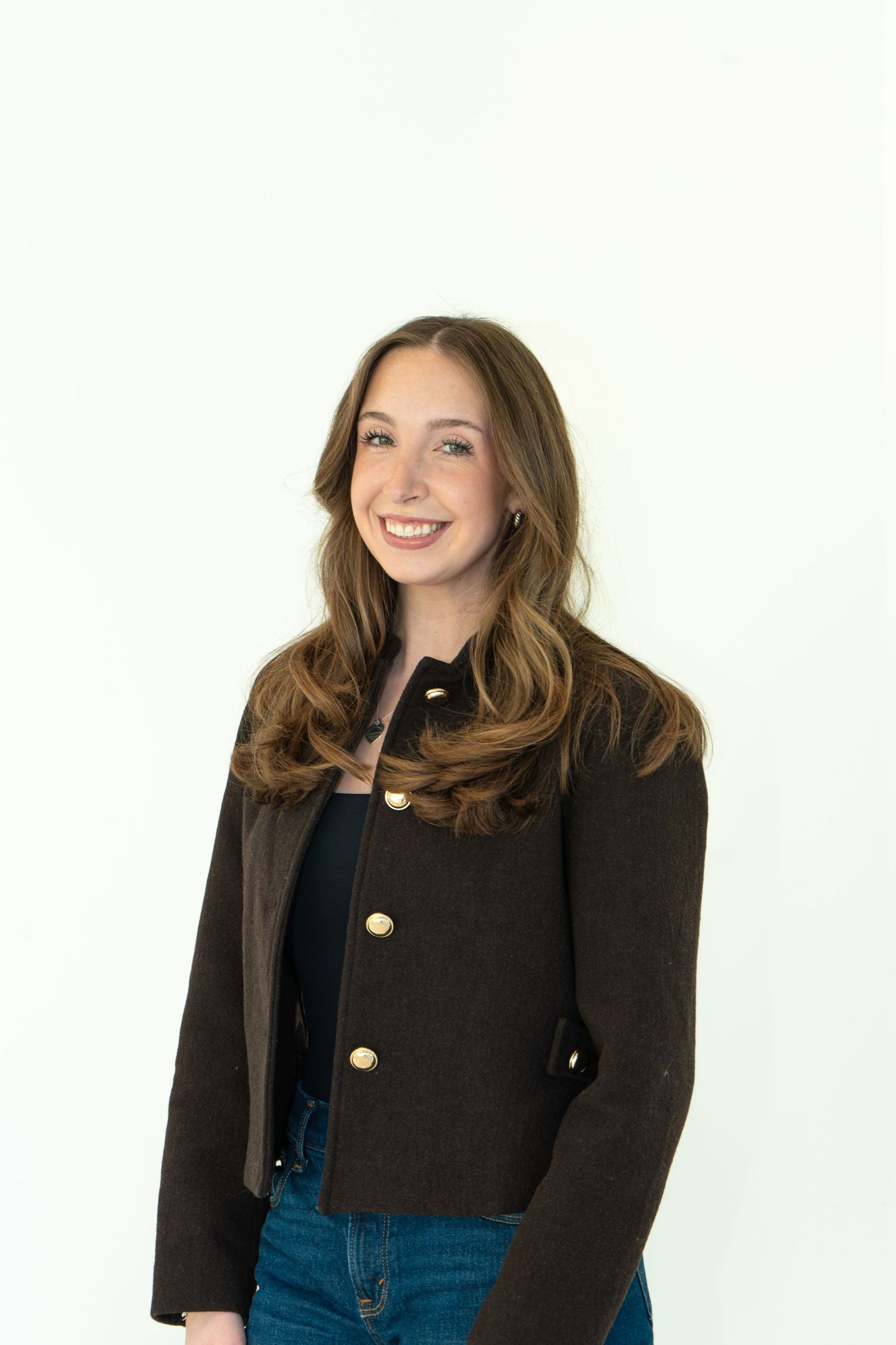 Smiling young woman with long brown hair wearing a dark blazer with gold buttons and blue jeans against white background.