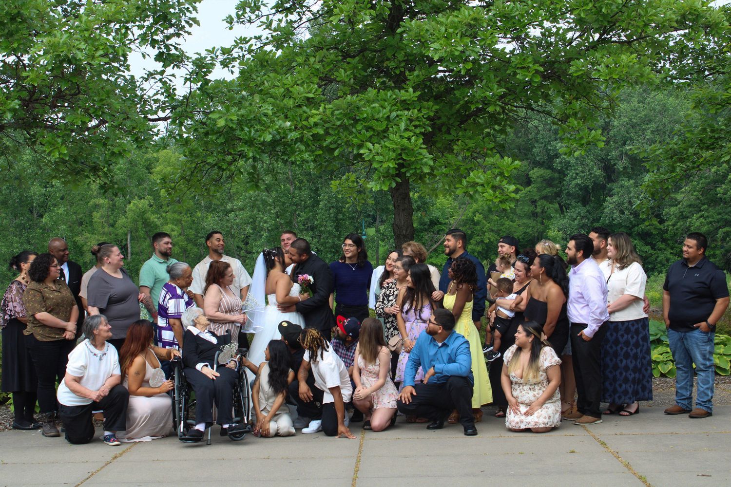 Large family group photo outdoors at a wedding celebration, posing together near lush green trees.