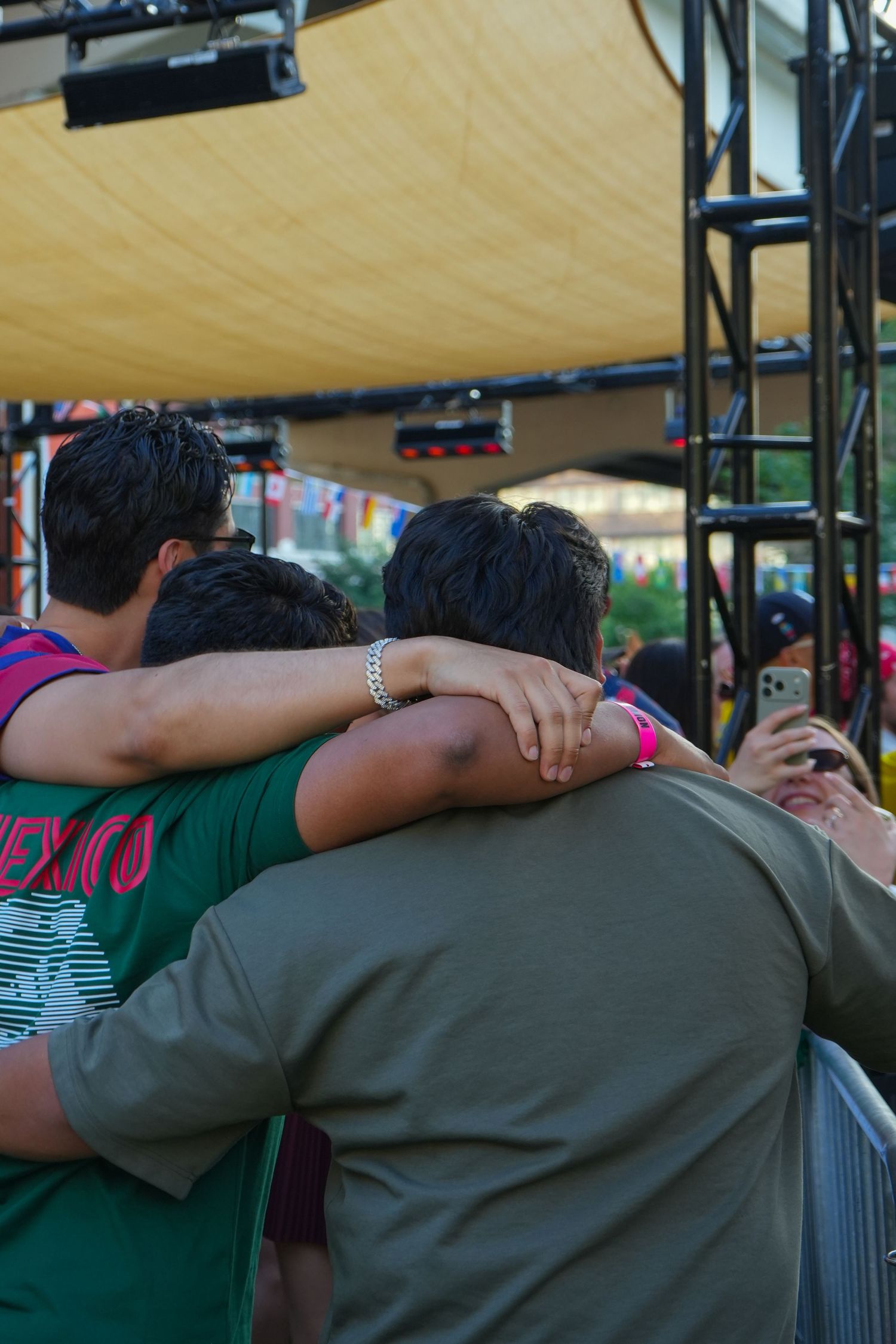 Three friends hugging each other from behind at an outdoor concert stage event, wearing casual shirts.