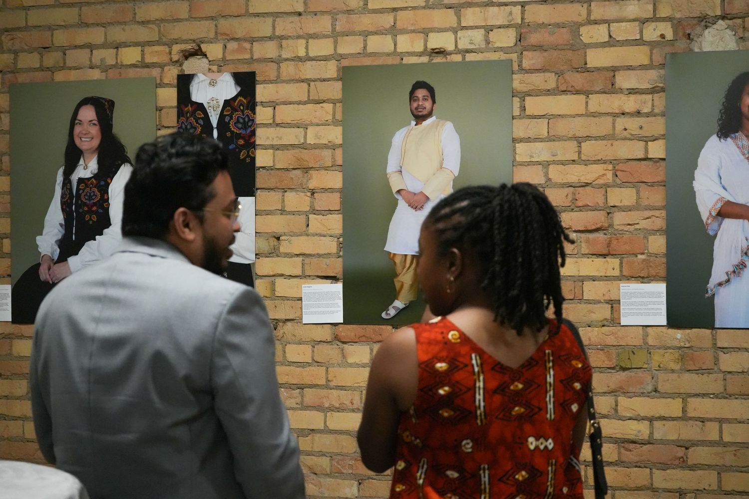 Two visitors viewing a multicultural photography exhibition displayed on a brick wall at an art gallery.