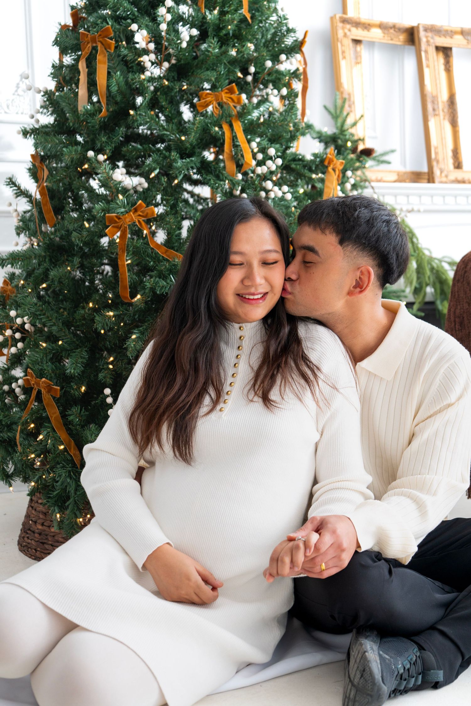 Pregnant couple sitting together by decorated Christmas tree, man kissing woman's cheek, both wearing white sweaters.
