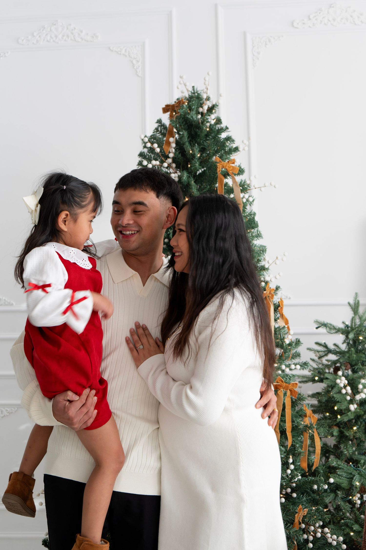 Happy family of three posing by decorated Christmas tree, parents holding young daughter in red dress in bright white room.
