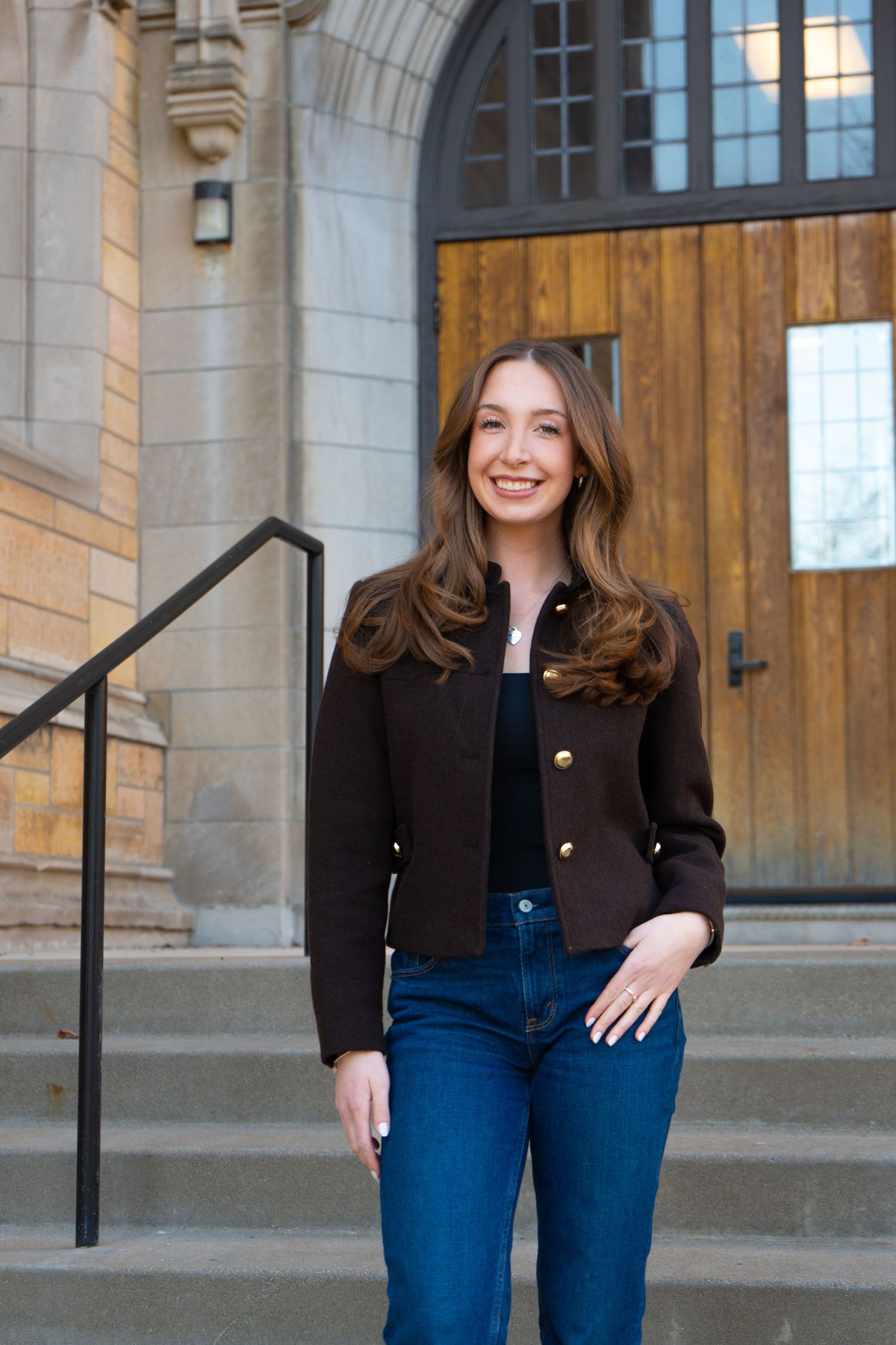 Young woman in black blazer and jeans smiling on steps of a stone building with wooden doors.
