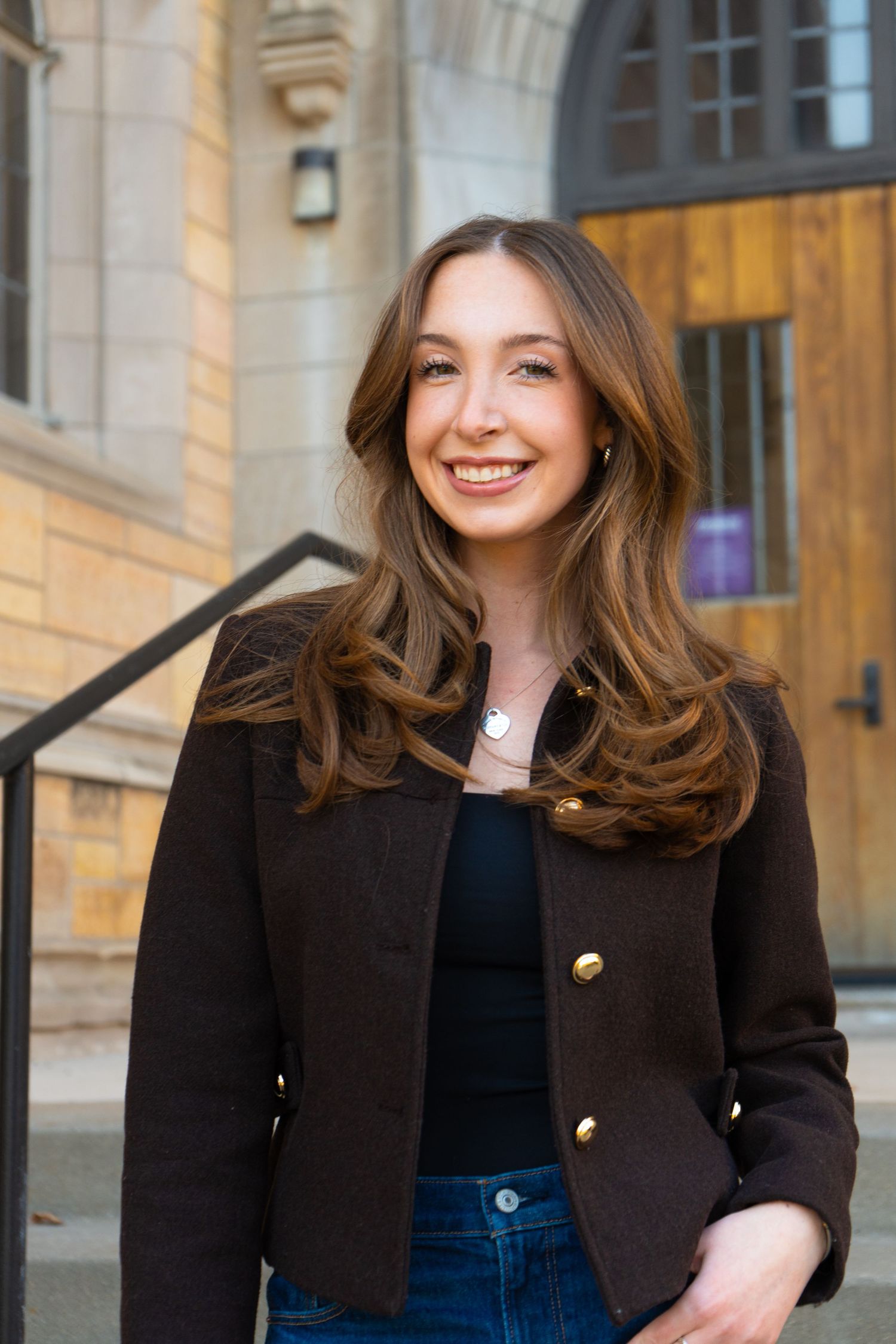 Smiling young woman with brown hair wearing a dark blazer with gold buttons, standing outside a stone building.