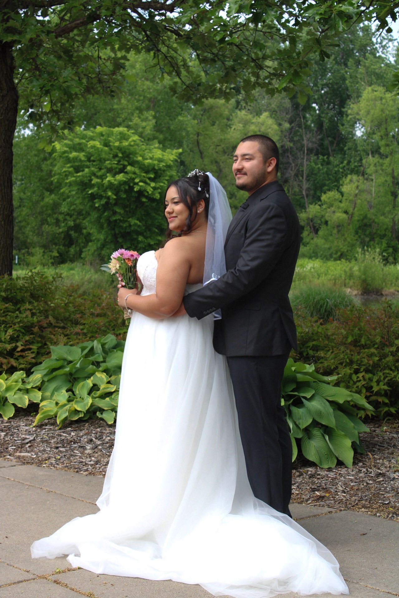 Bride in white gown holding bouquet stands with groom in black suit outdoors in lush green garden setting.