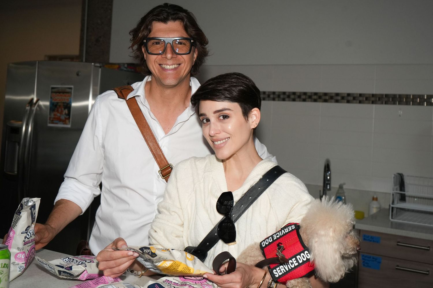 Two smiling people pose together indoors, with a service dog vest visible in the foreground.