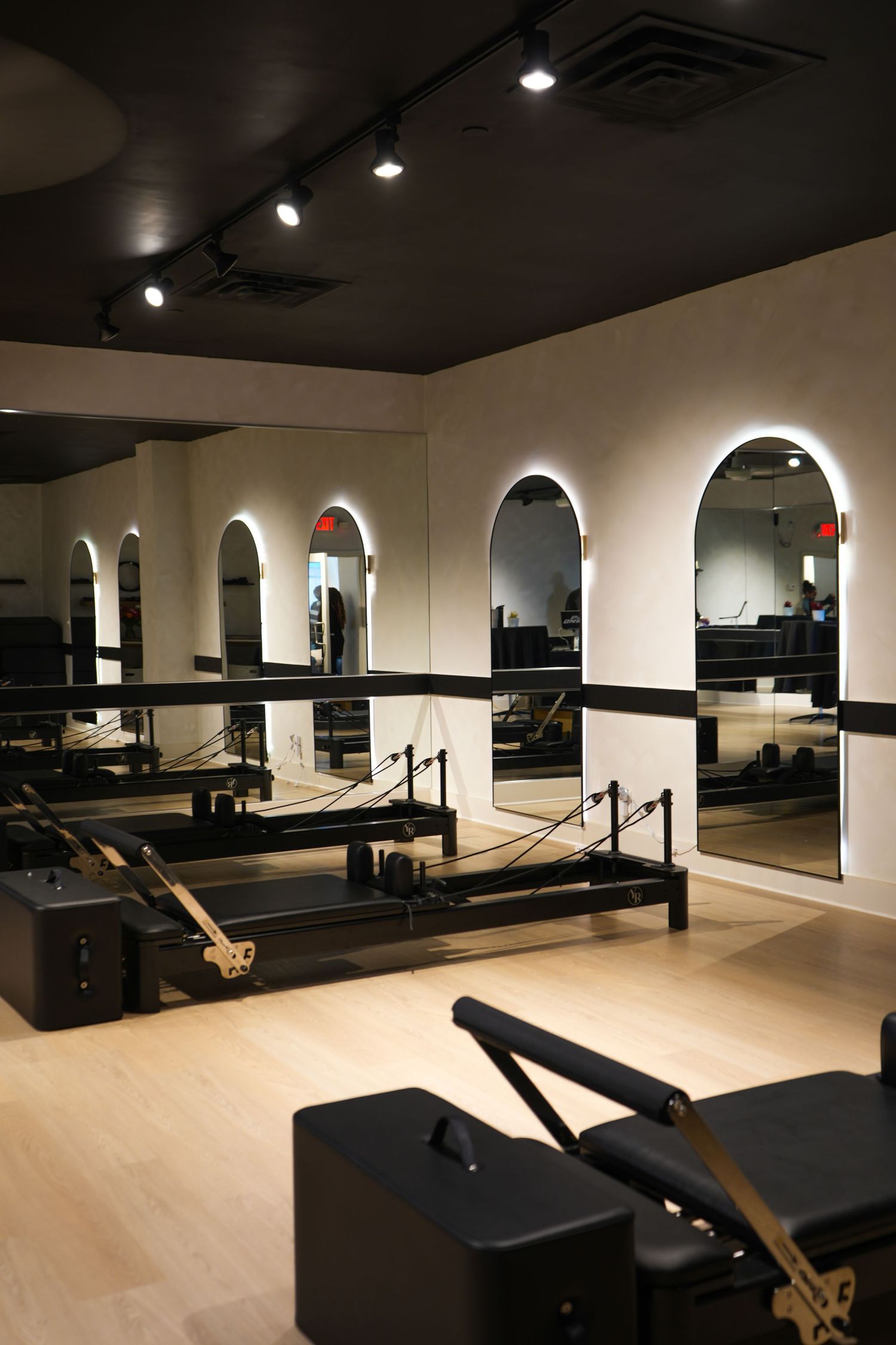 Modern Pilates studio with reformer machines, arched mirrors, and track lighting on hardwood floors.