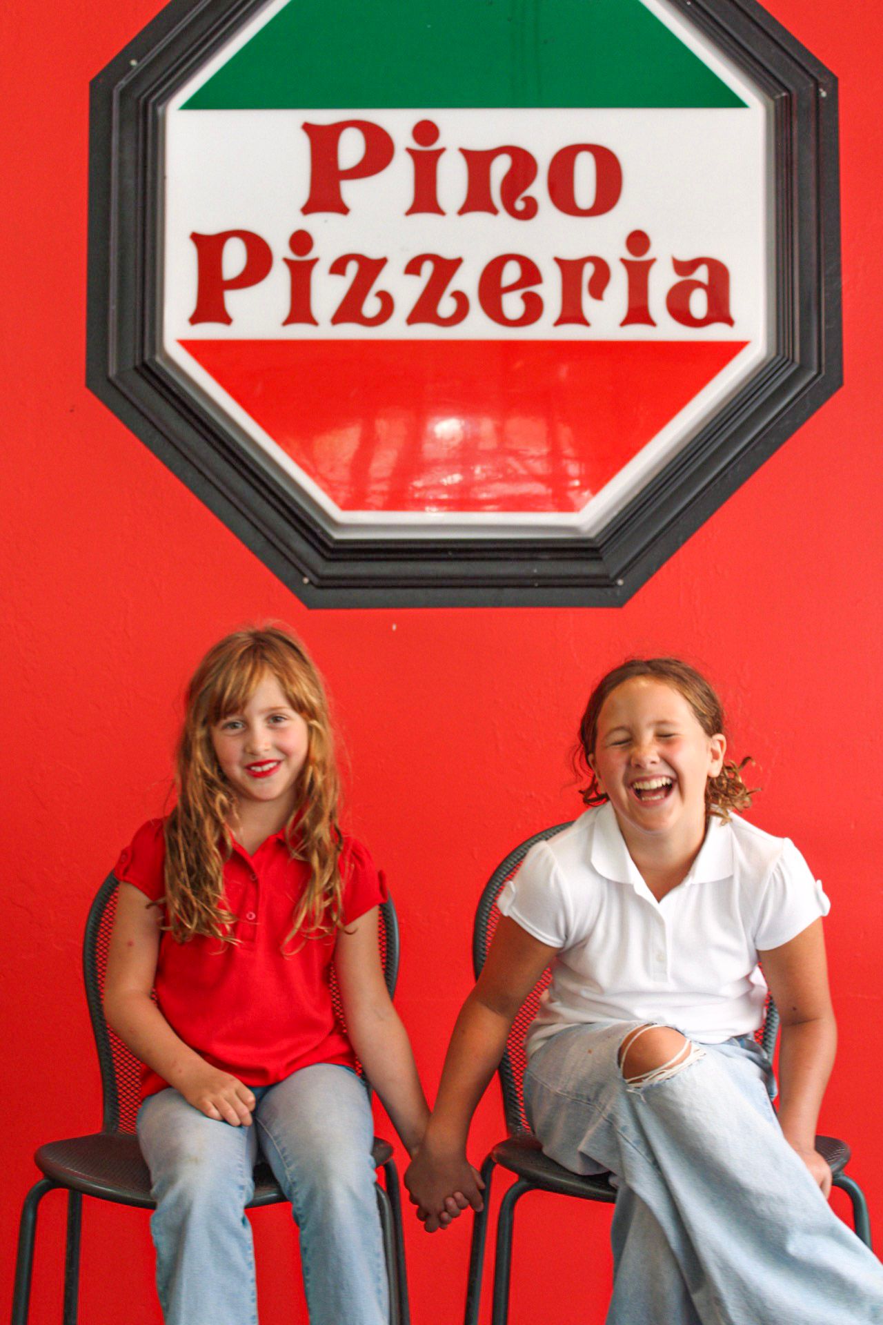 Two young girls laughing and holding hands outside Pino Pizzeria restaurant with red wall backdrop.