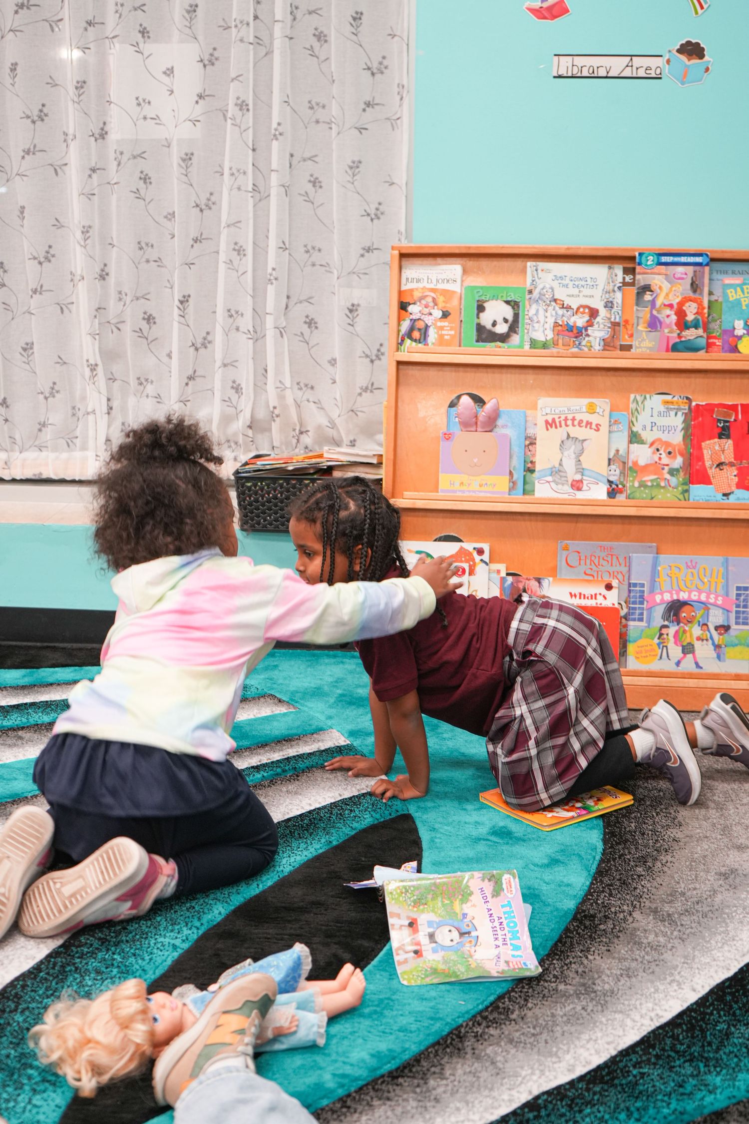 Two young girls playing together on a colorful rug in a classroom library area with bookshelves in background.