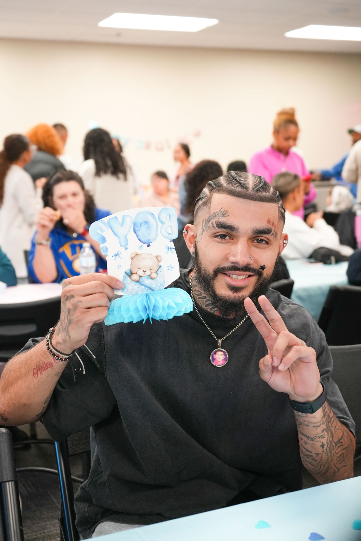 Tattooed man smiling at a baby shower, holding a blue boy decoration and flashing a peace sign.