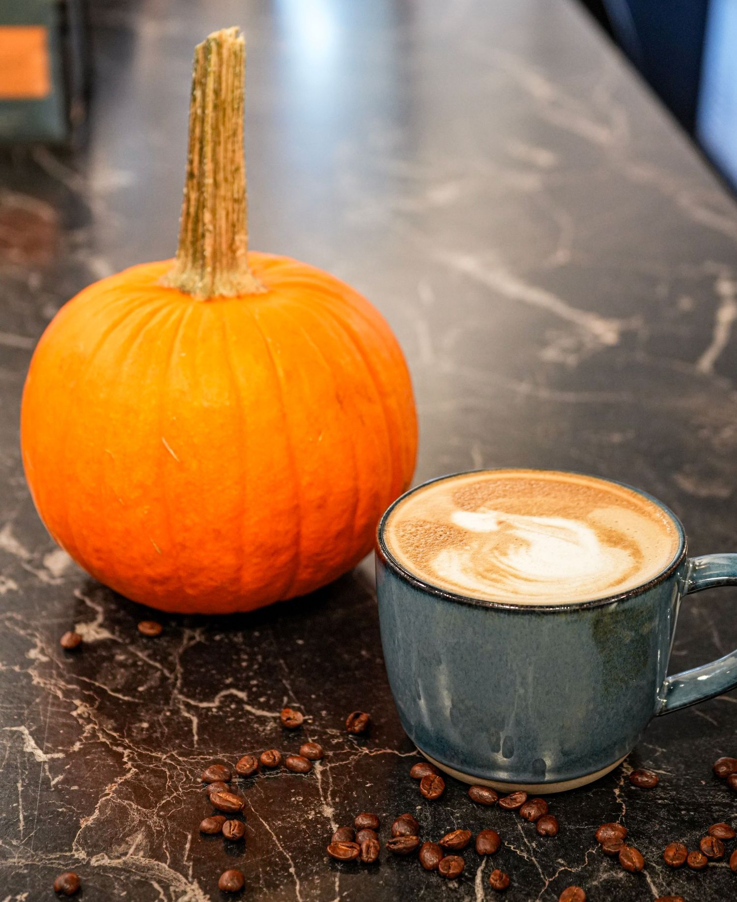 Orange pumpkin beside a blue ceramic mug of latte art on dark marble surface with scattered coffee beans.