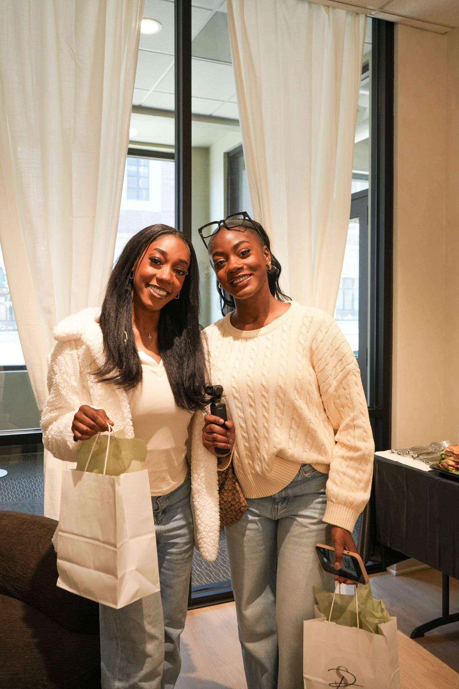 Two smiling women in cream sweaters and jeans pose together holding gift bags indoors.