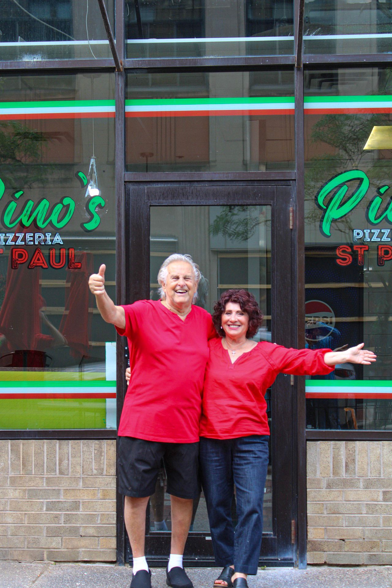 Two smiling owners in red shirts pose proudly outside Pino's Pizzeria in St. Paul, giving thumbs up.
