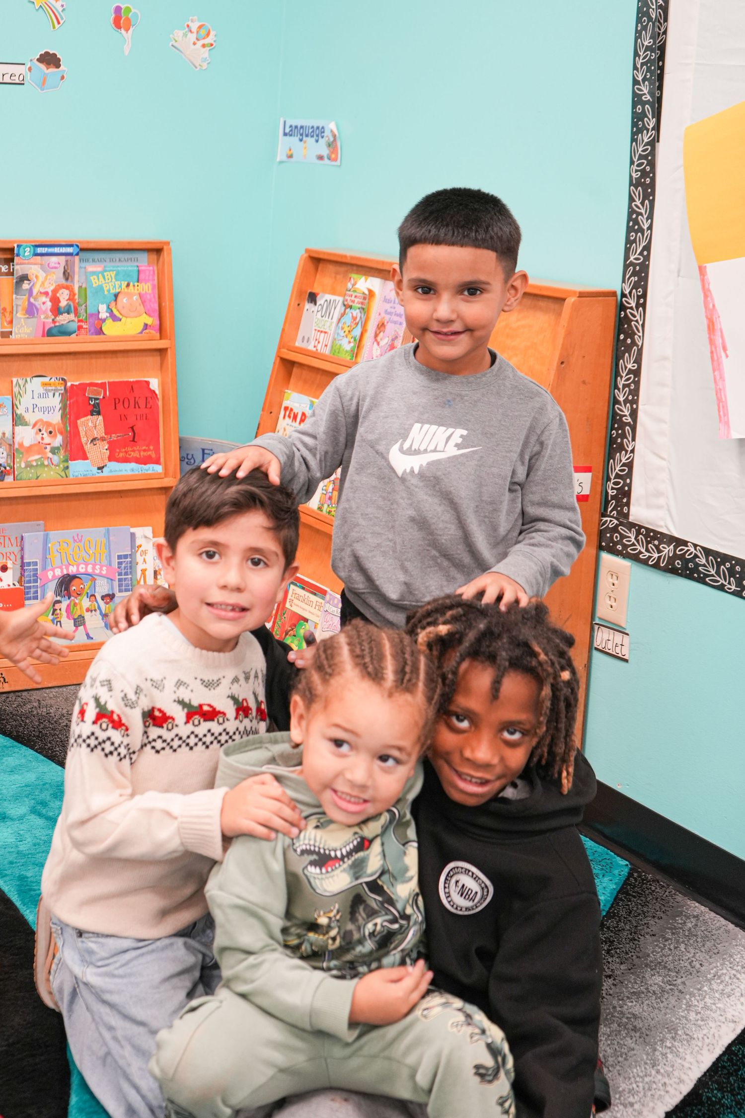 Four young children smile together in a colorful classroom library with bookshelves in the background.