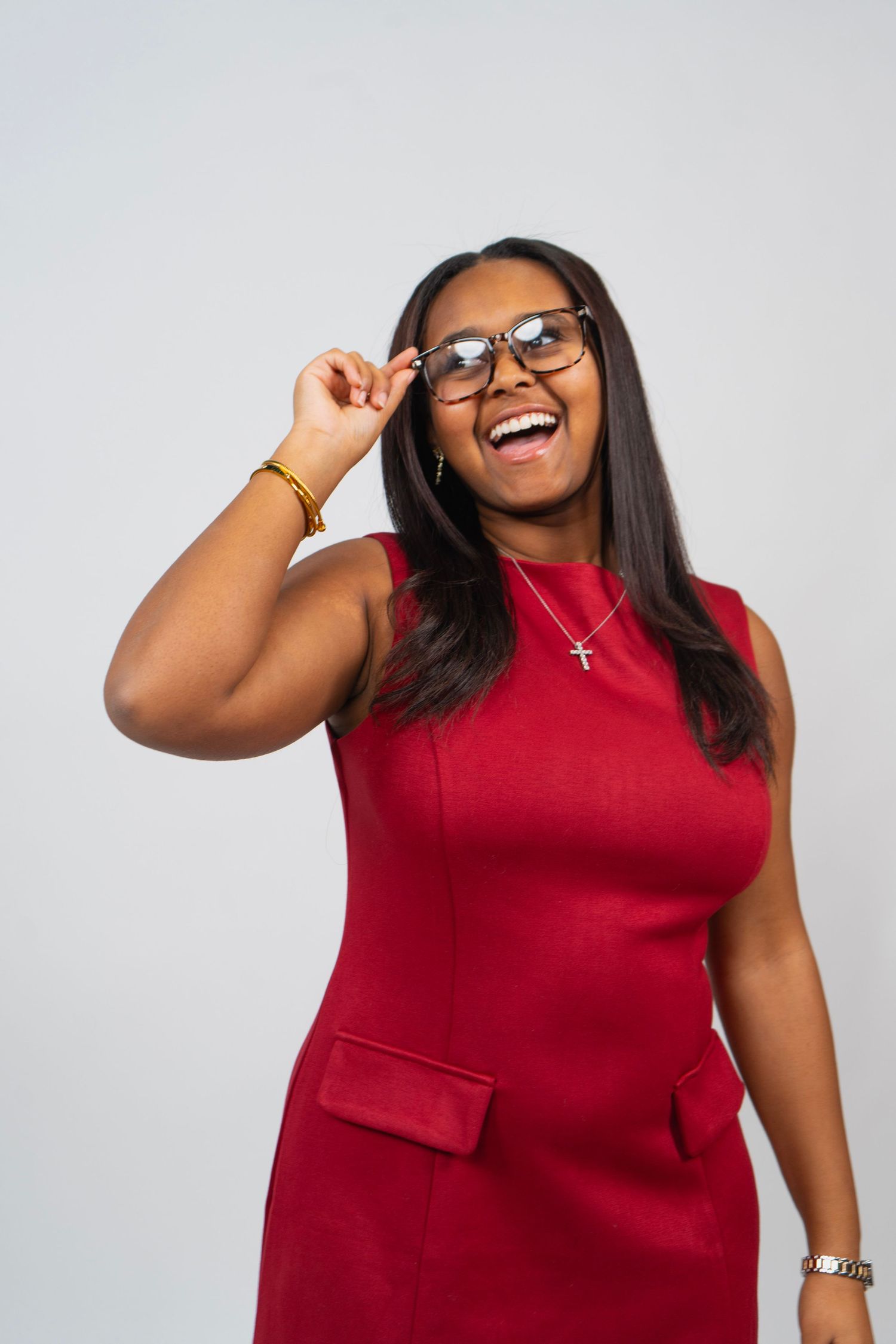 Smiling Black woman in red dress adjusting glasses, wearing cross necklace, posing against gray background.