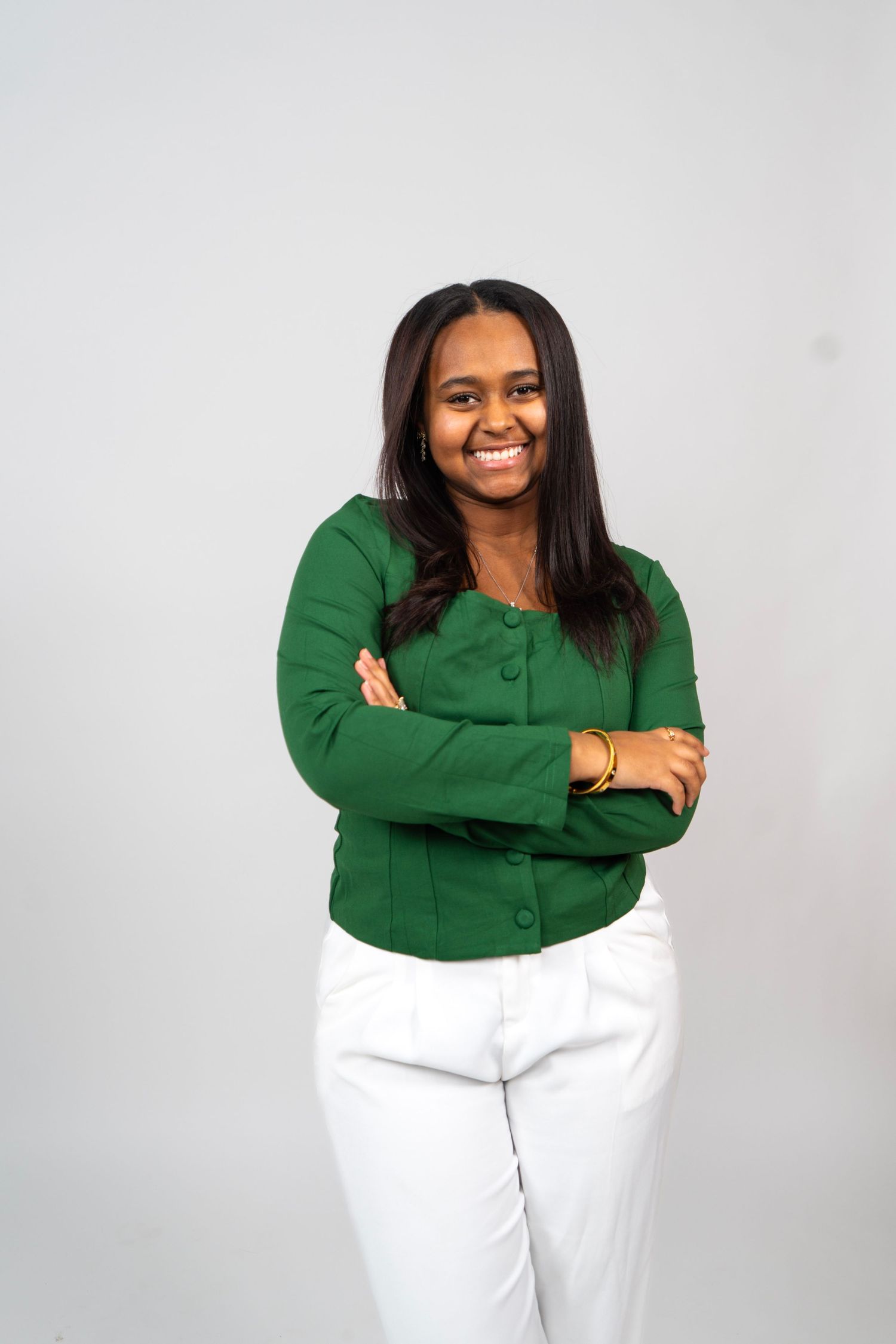 Smiling young Black woman in green cardigan and white pants standing with arms crossed against gray background.