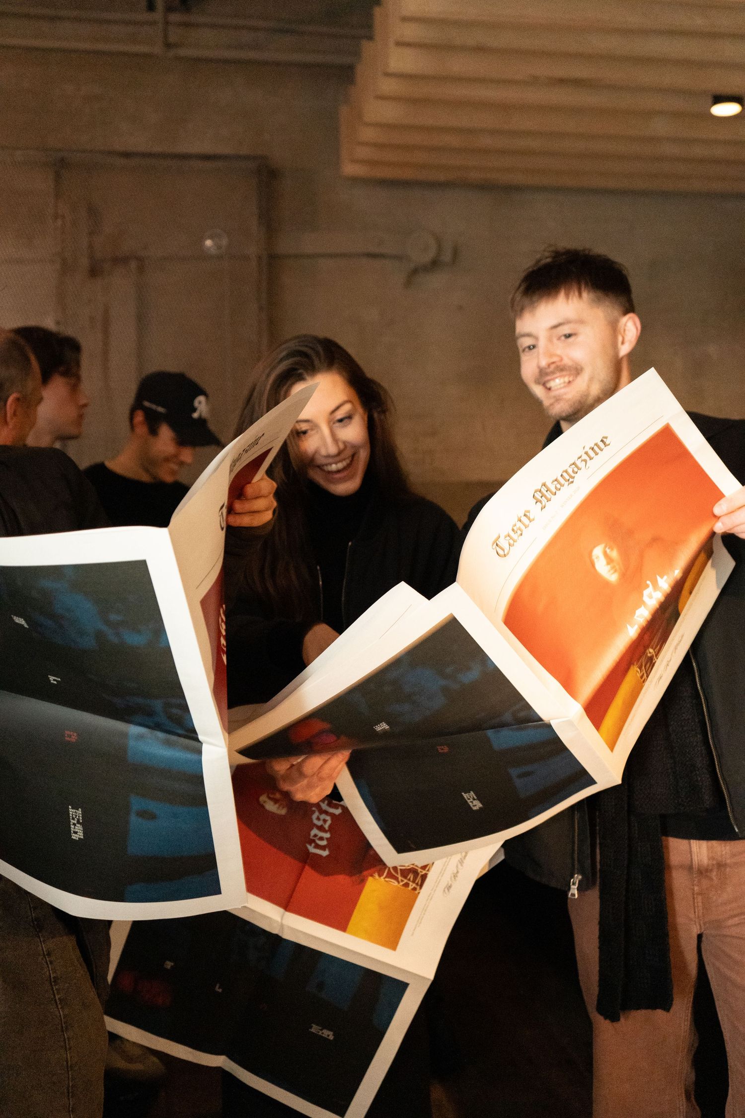Two people laughing while reading a large print magazine at an indoor event.