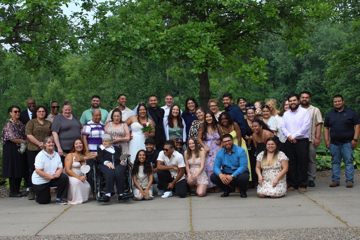 Large family group photo outdoors with lush green trees, featuring adults and children posing together at a formal gathering.
