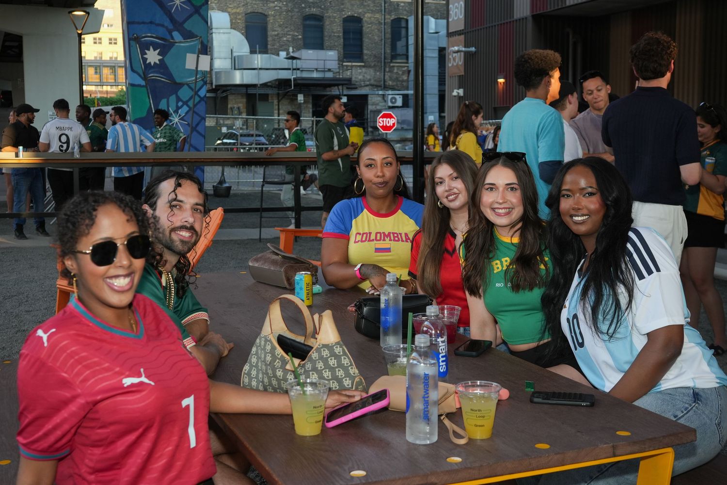 Group of friends in sports jerseys smiling at outdoor bar table during soccer World Cup event.