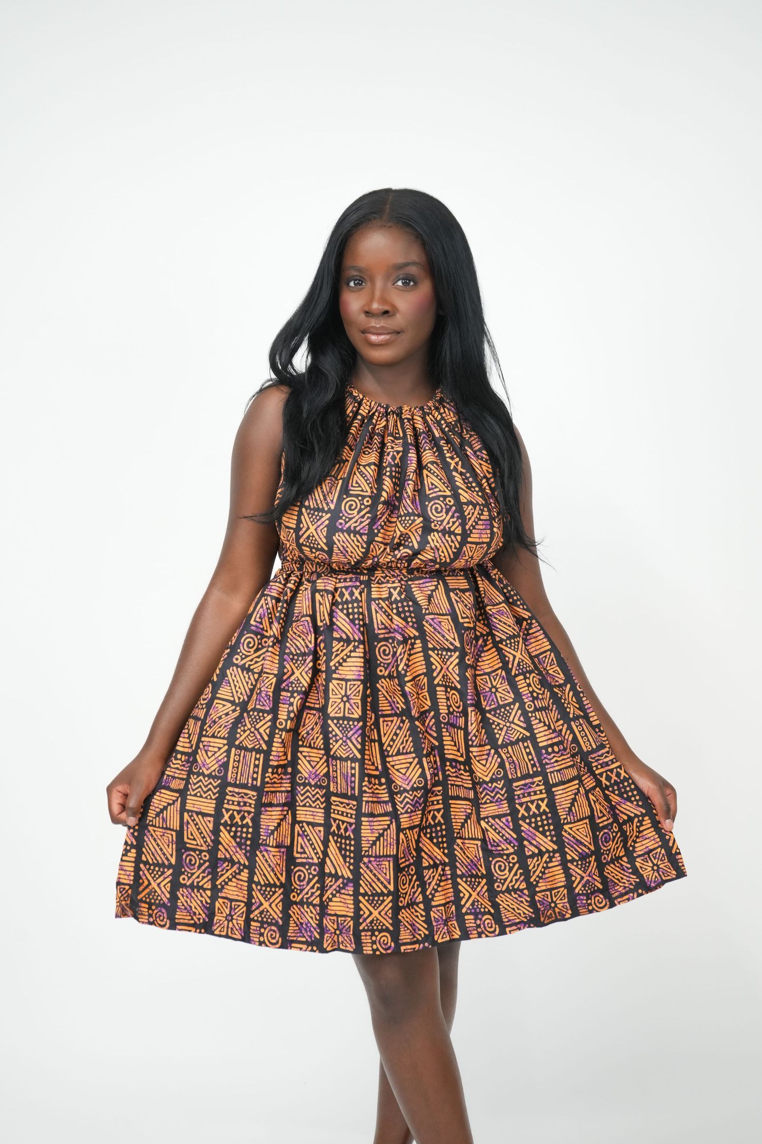 Young Black woman wearing a vibrant African print halter dress with geometric patterns, posing against a white background.