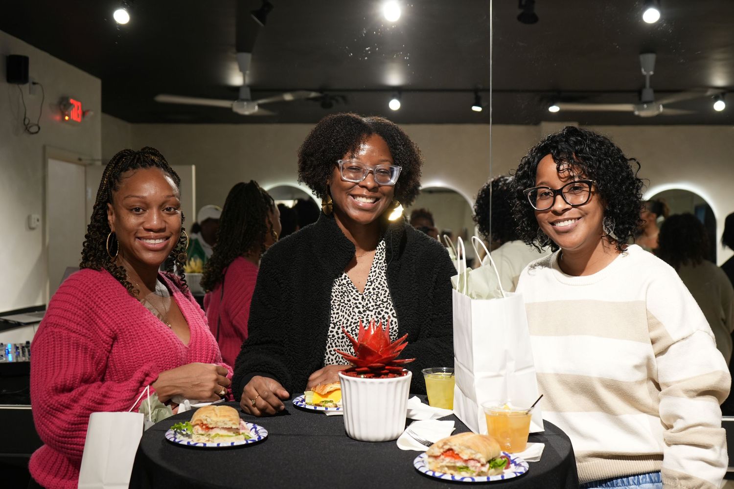 Three women smiling together at a round dining table with food and drinks at an indoor social gathering event.