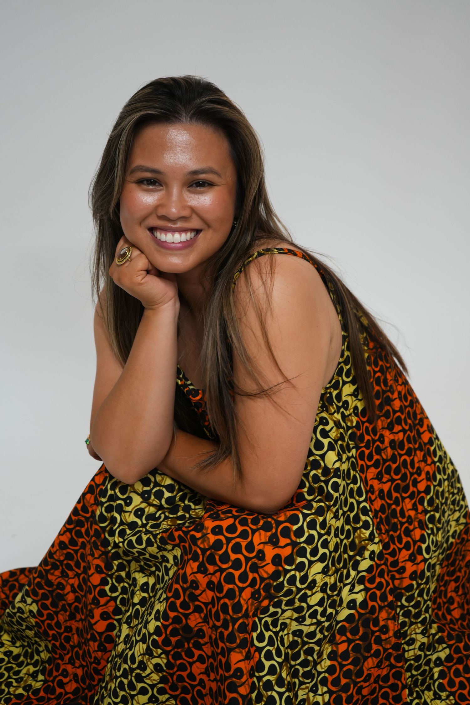 Smiling young woman in colorful orange and green patterned dress, posing with hand on chin against gray background.