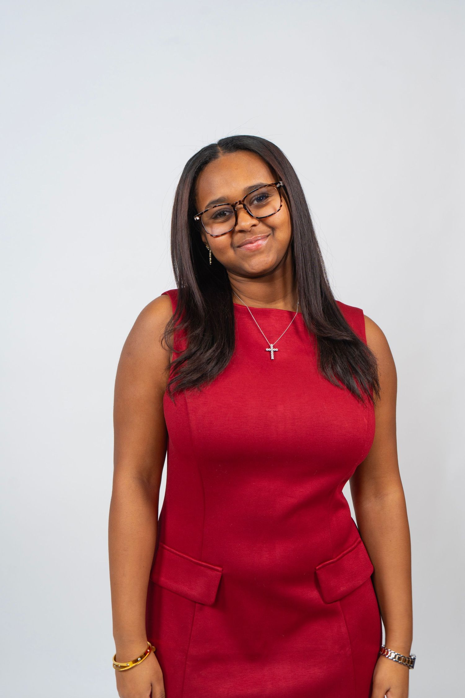 Young woman in red sleeveless dress and glasses wearing a cross necklace, smiling against a light gray background.
