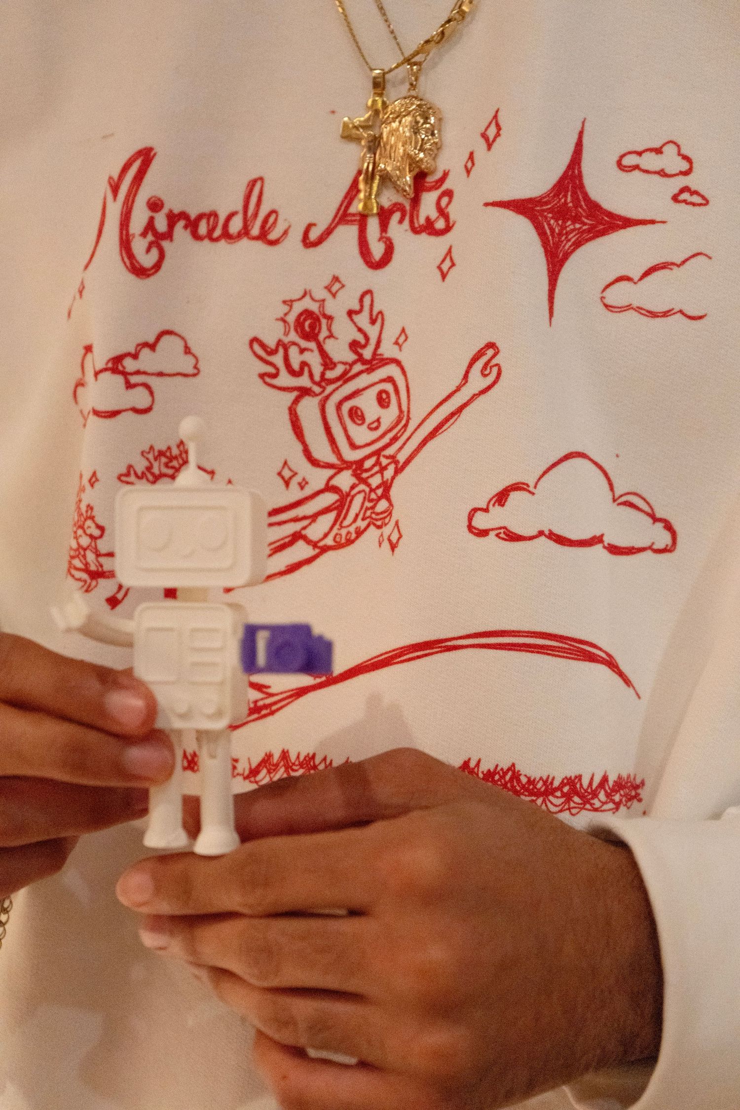 Person holding a white robot toy figurine, wearing a Miracle Arts graphic sweatshirt with red illustrations and gold necklace.