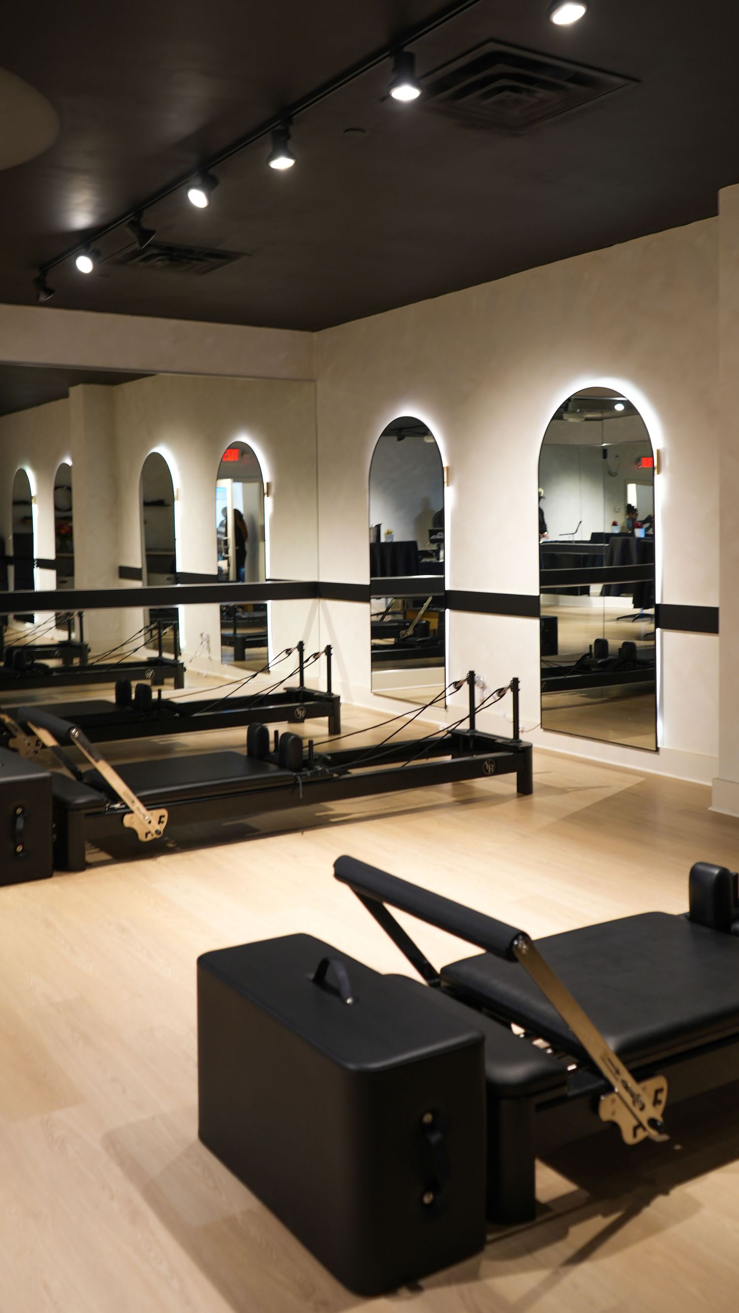 Modern Pilates studio with black reformer machines, arched mirrors, and track lighting on light wood floors.