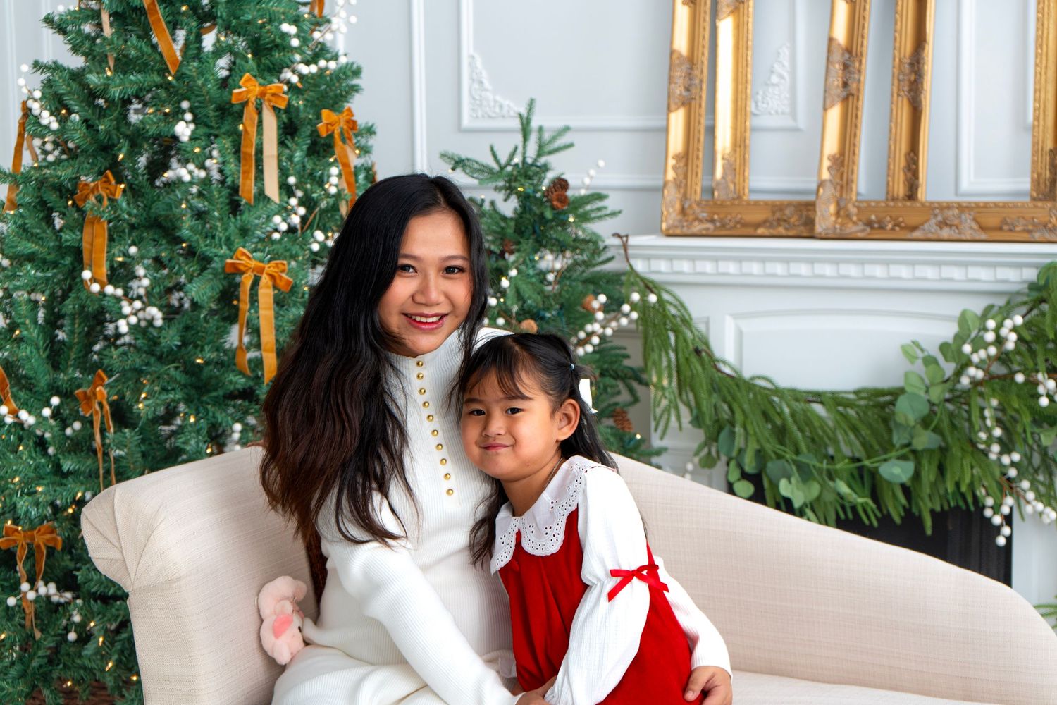 Woman and young girl in festive outfits smiling together on a sofa beside a decorated Christmas tree.