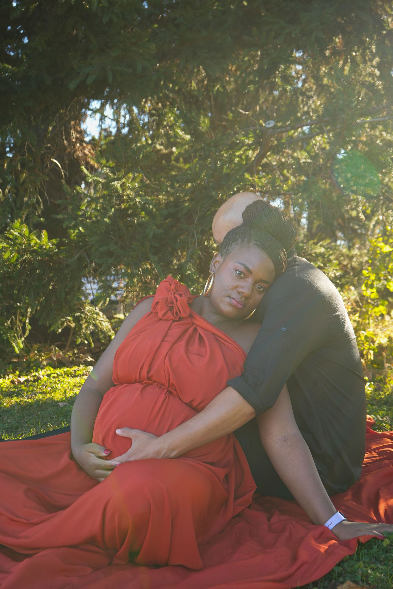 Pregnant woman in red gown sits outdoors with partner embracing her from behind, golden sunlight filtering through green trees.